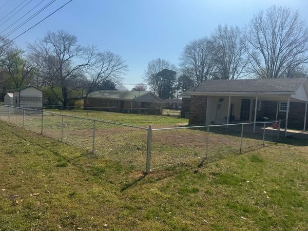 A fenced-in yard with dried grass in front of several single-story residential houses under a clear blue sky.