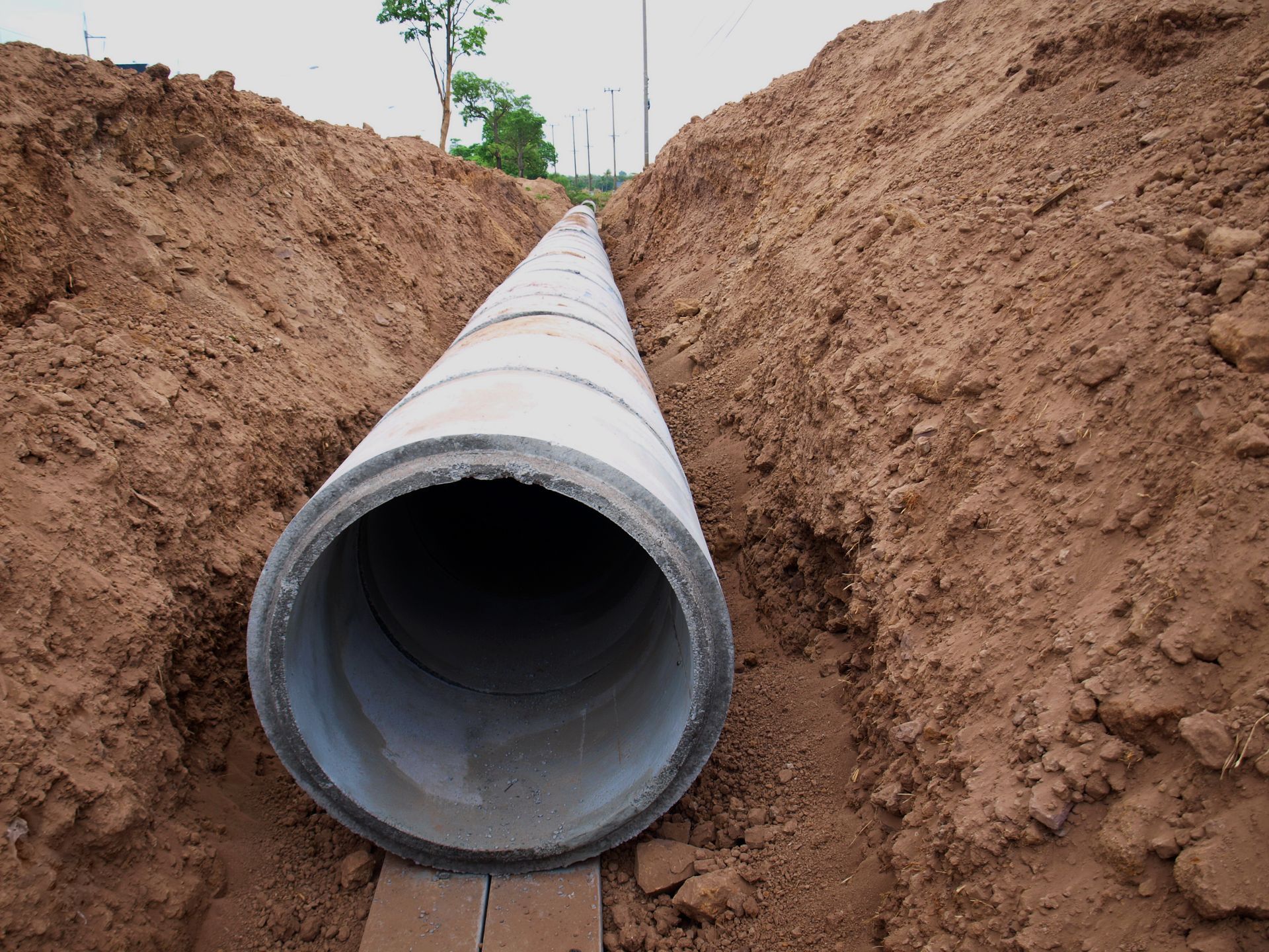 A large grey concrete pipe installed in a deep, narrow dirt trench at a construction site.