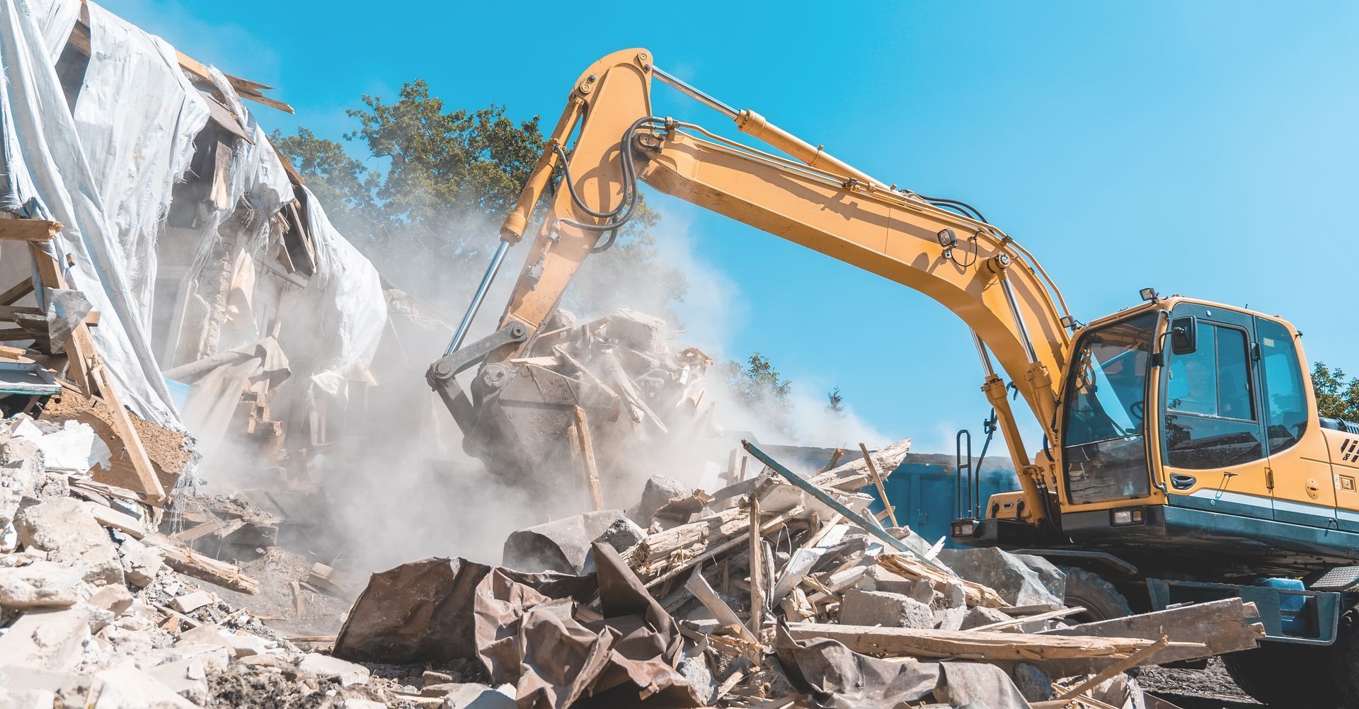 A yellow excavator demolishing a building, kicking up a cloud of dust against a clear blue sky.