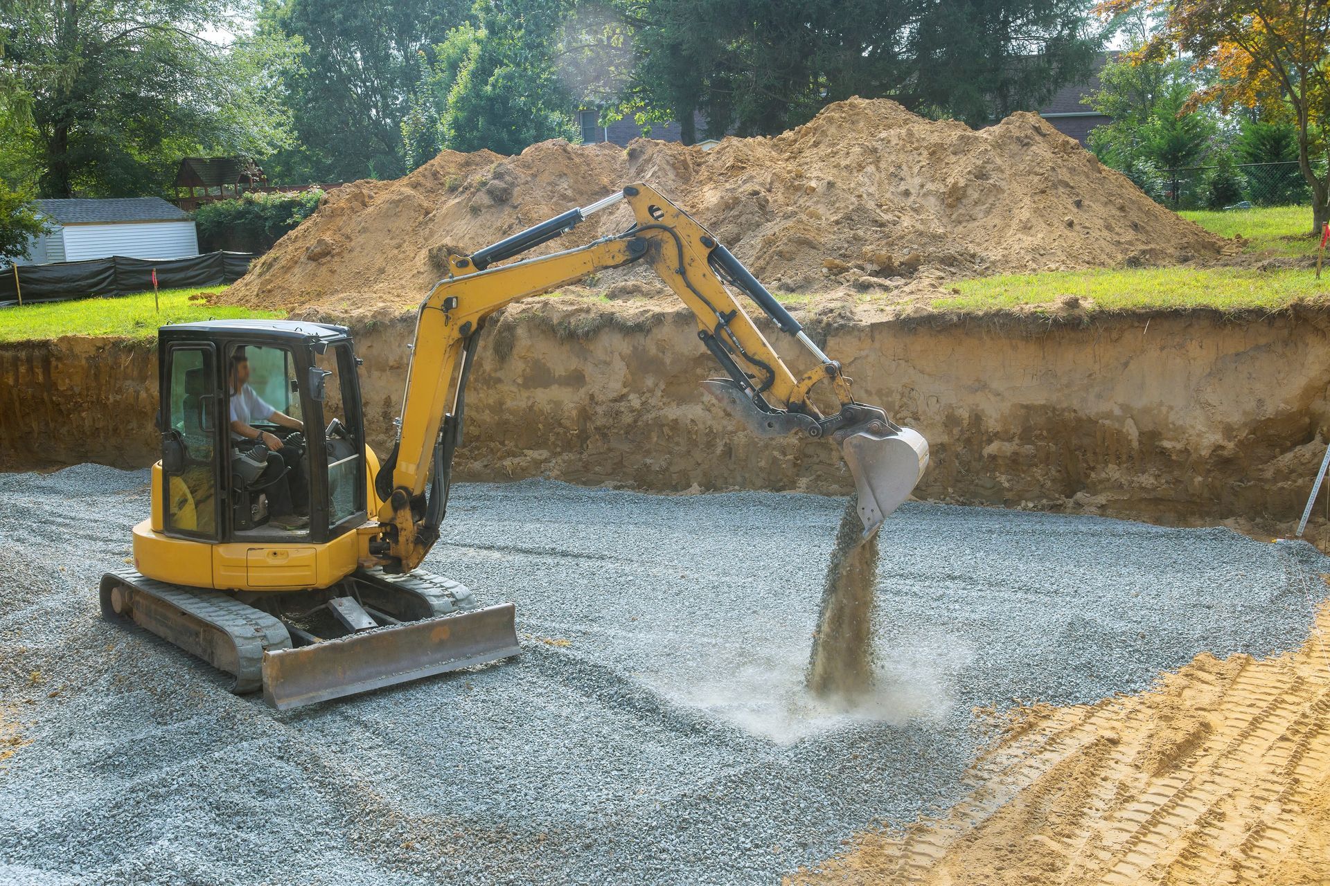 A yellow mini excavator spreads gravel inside a deep excavated foundation pit on a construction site.