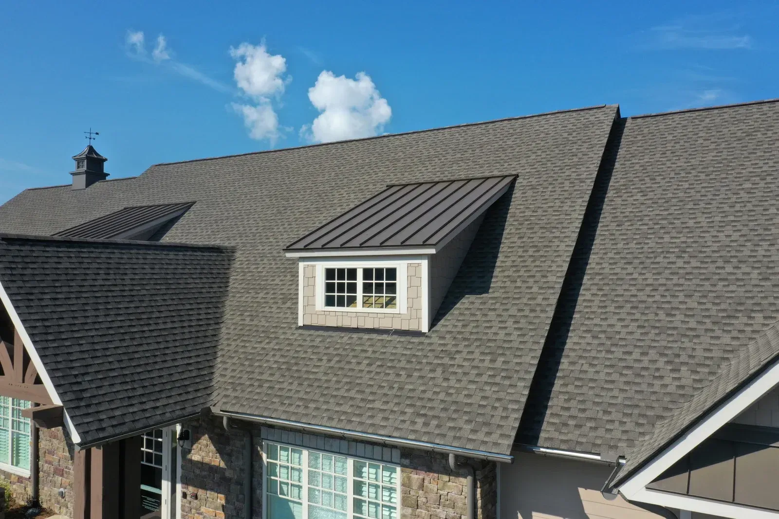 Gray asphalt shingle roof on a house, dormer with window. Blue sky and white clouds in background.