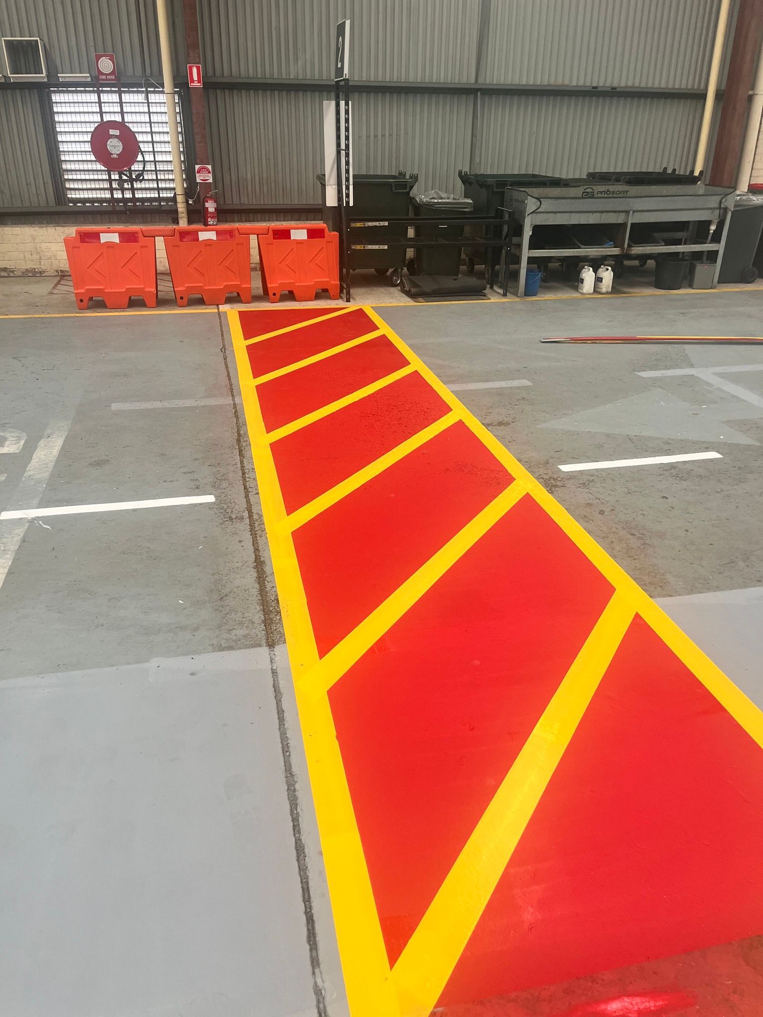 Red and yellow painted pathway with diagonal stripes, leading to orange barriers in an industrial setting.