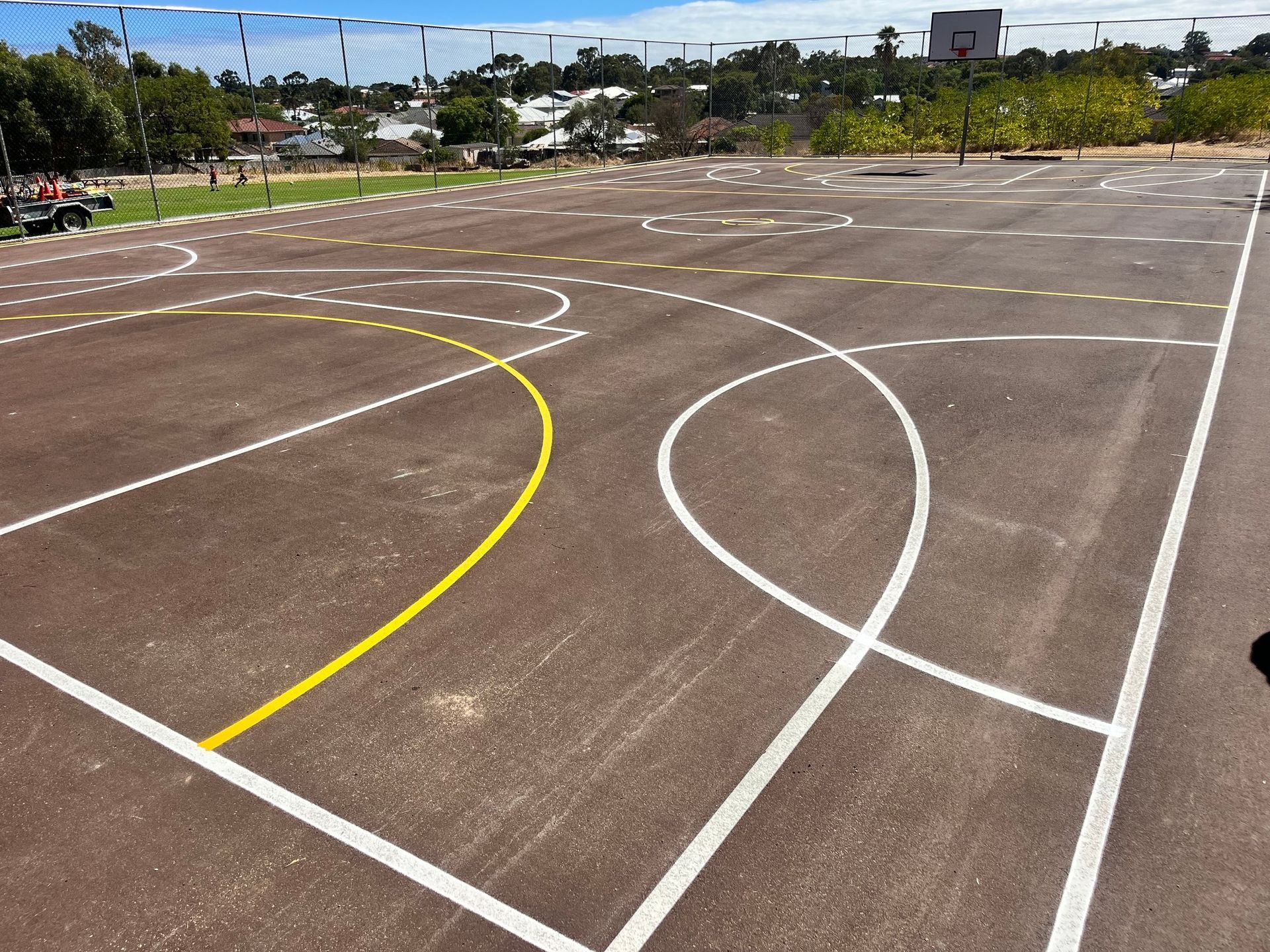 Multi-sport court with white and yellow lines, a volleyball net, and a backdrop of buildings and trees.