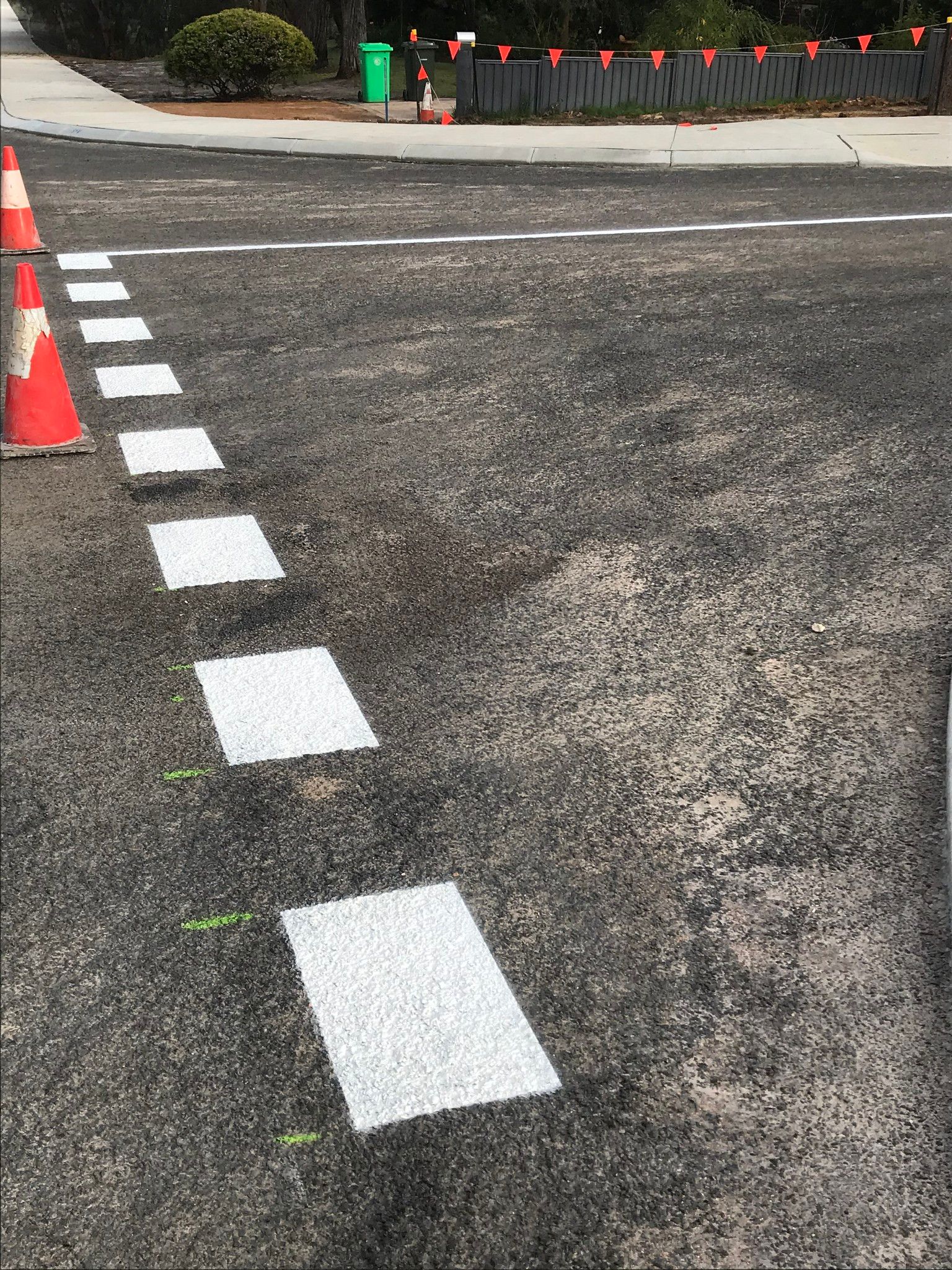 Asphalt road with white rectangular markings, orange cones, and a green trash can.