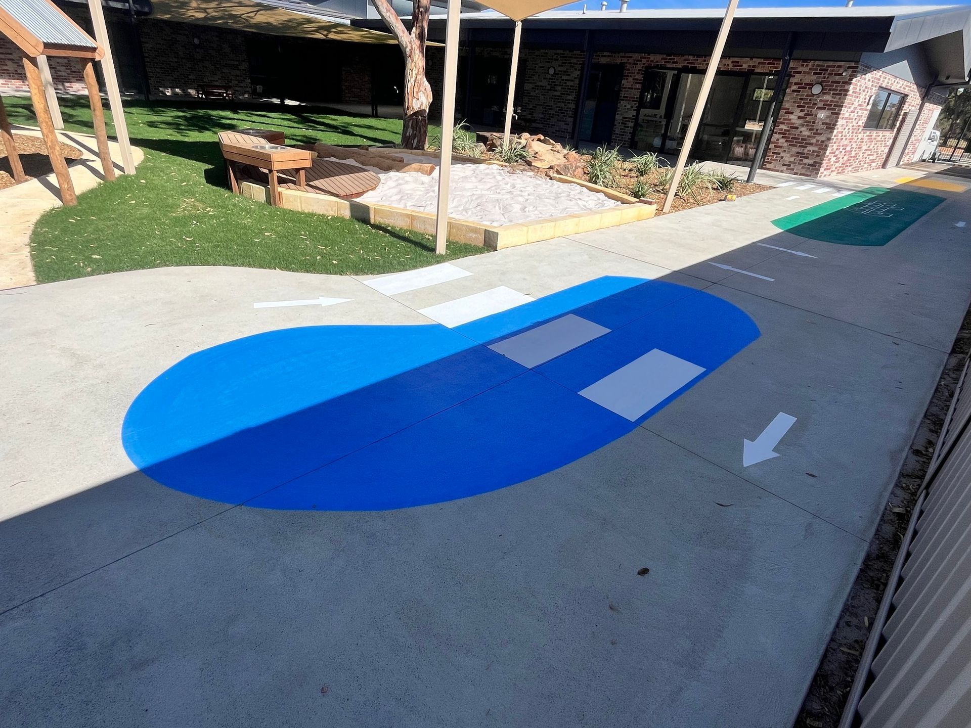 Playground with blue painted area and white markings. Concrete ground, green grass, and building in background.