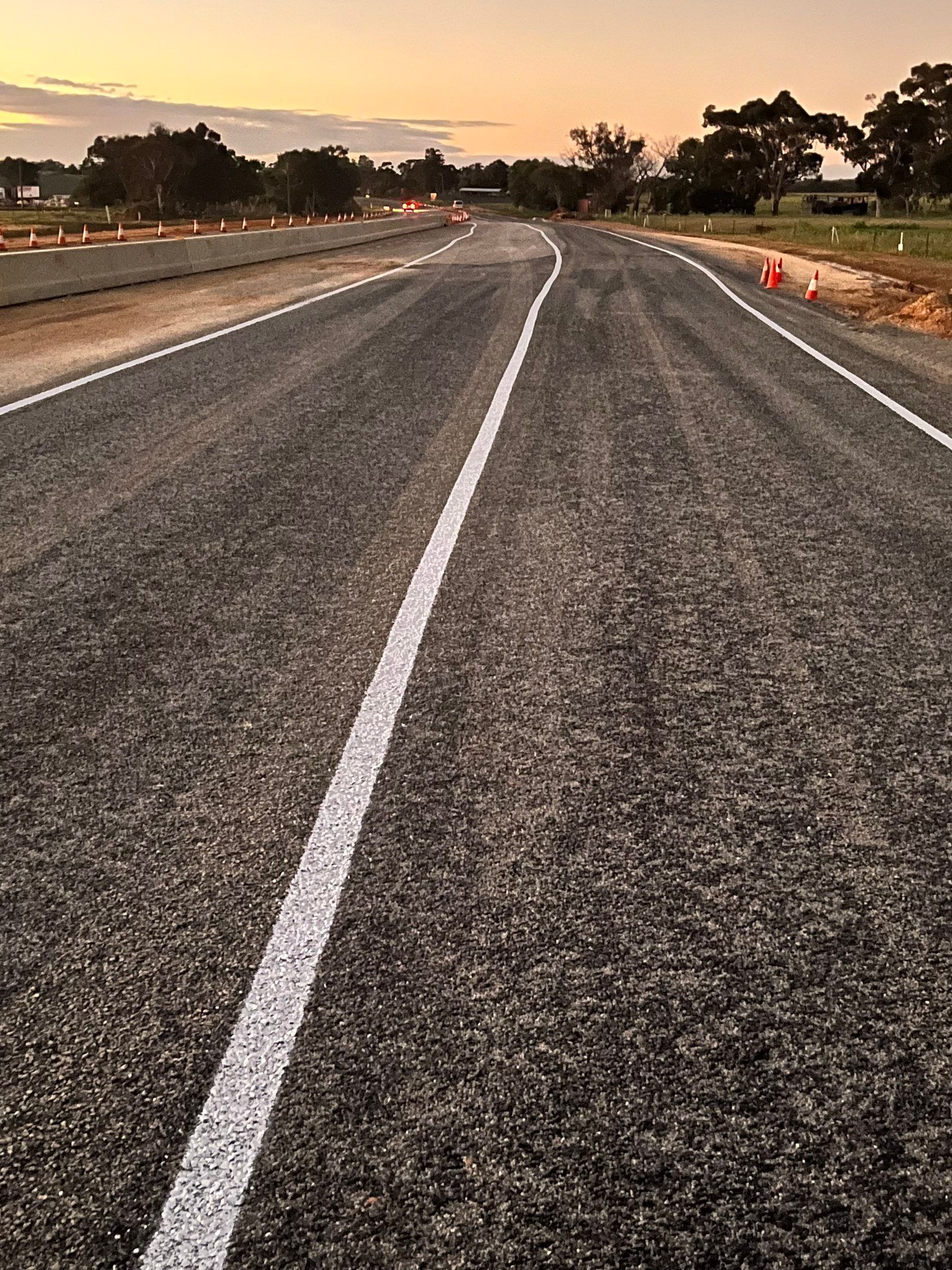 Asphalt road with white lines, trees on either side, orange cones, sunset.
