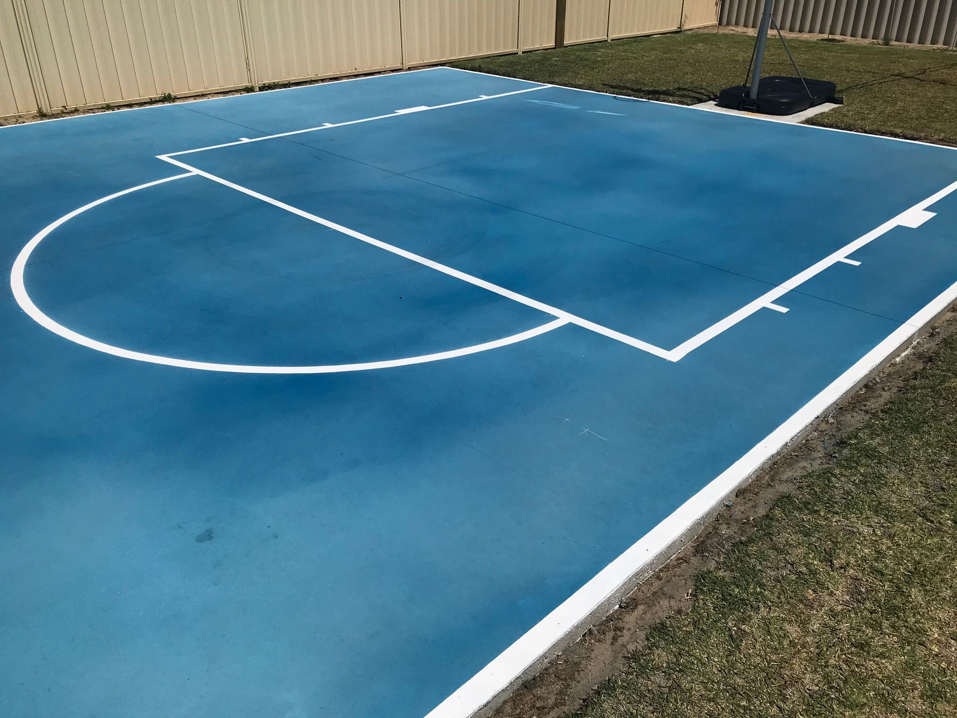 Blue outdoor basketball court with white lines and a tree in the background.