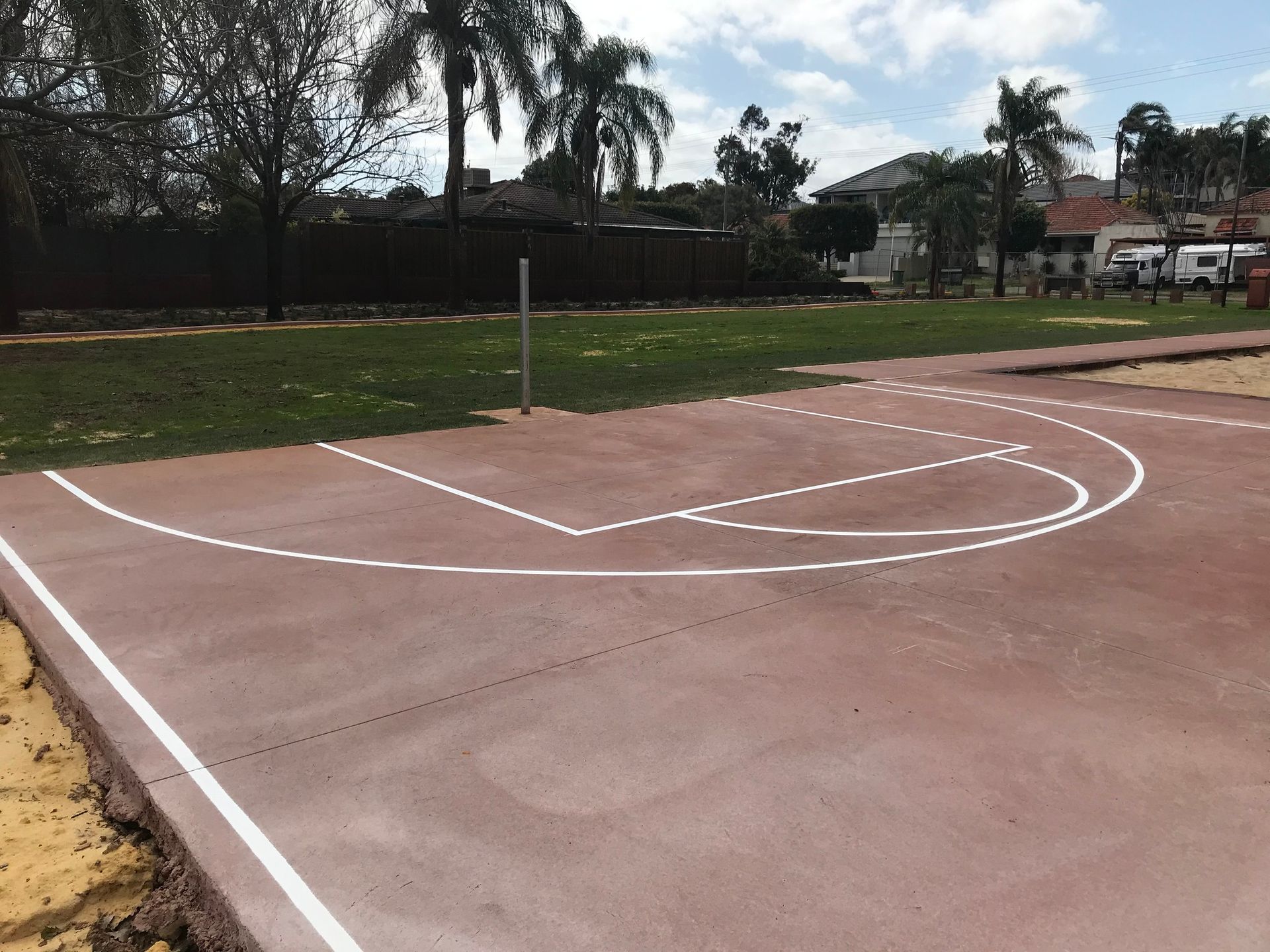 Outdoor basketball court with red surface and white lines, surrounded by grass and trees.