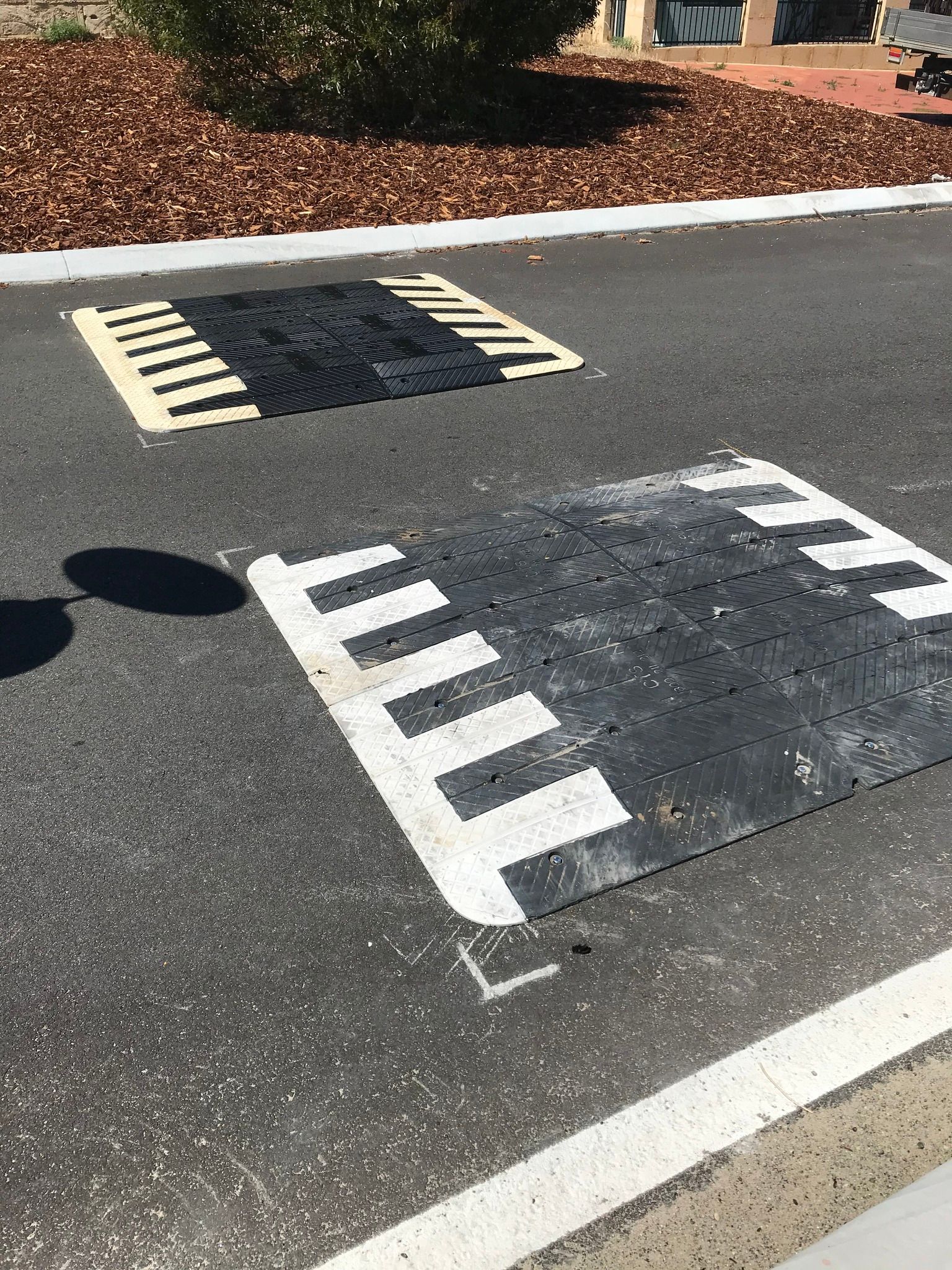Two black and white speed bumps on asphalt road, near a patch of mulch.