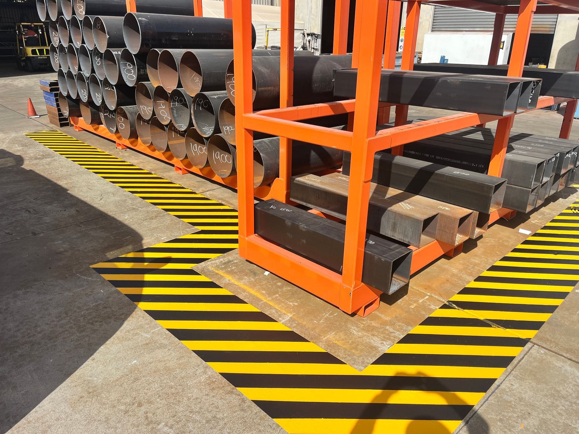 Orange metal shelving unit holding black pipes and square metal stock, on yellow and black striped floor markings.