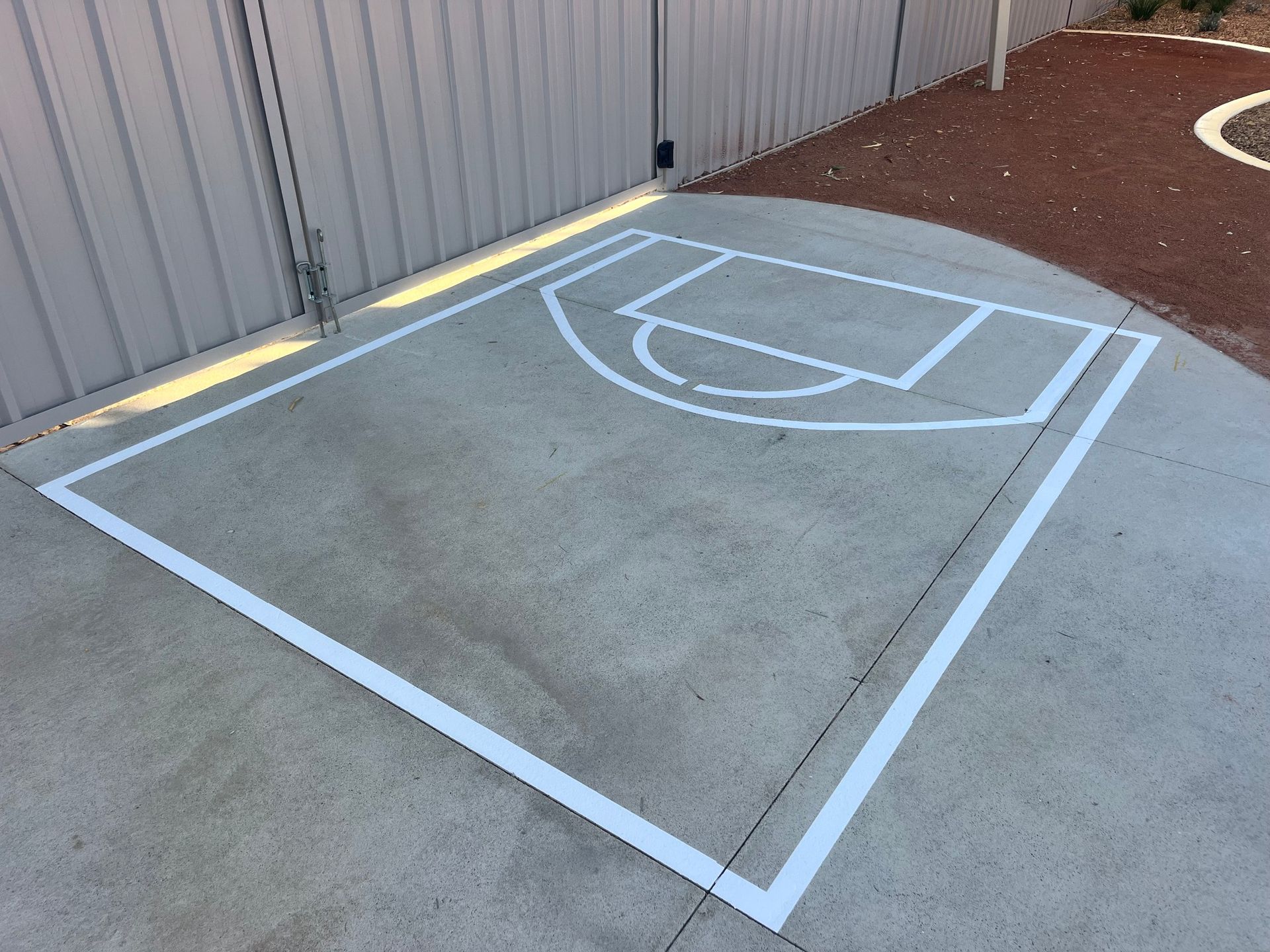 White-painted basketball court lines on a concrete surface next to a fence and a gravel area.