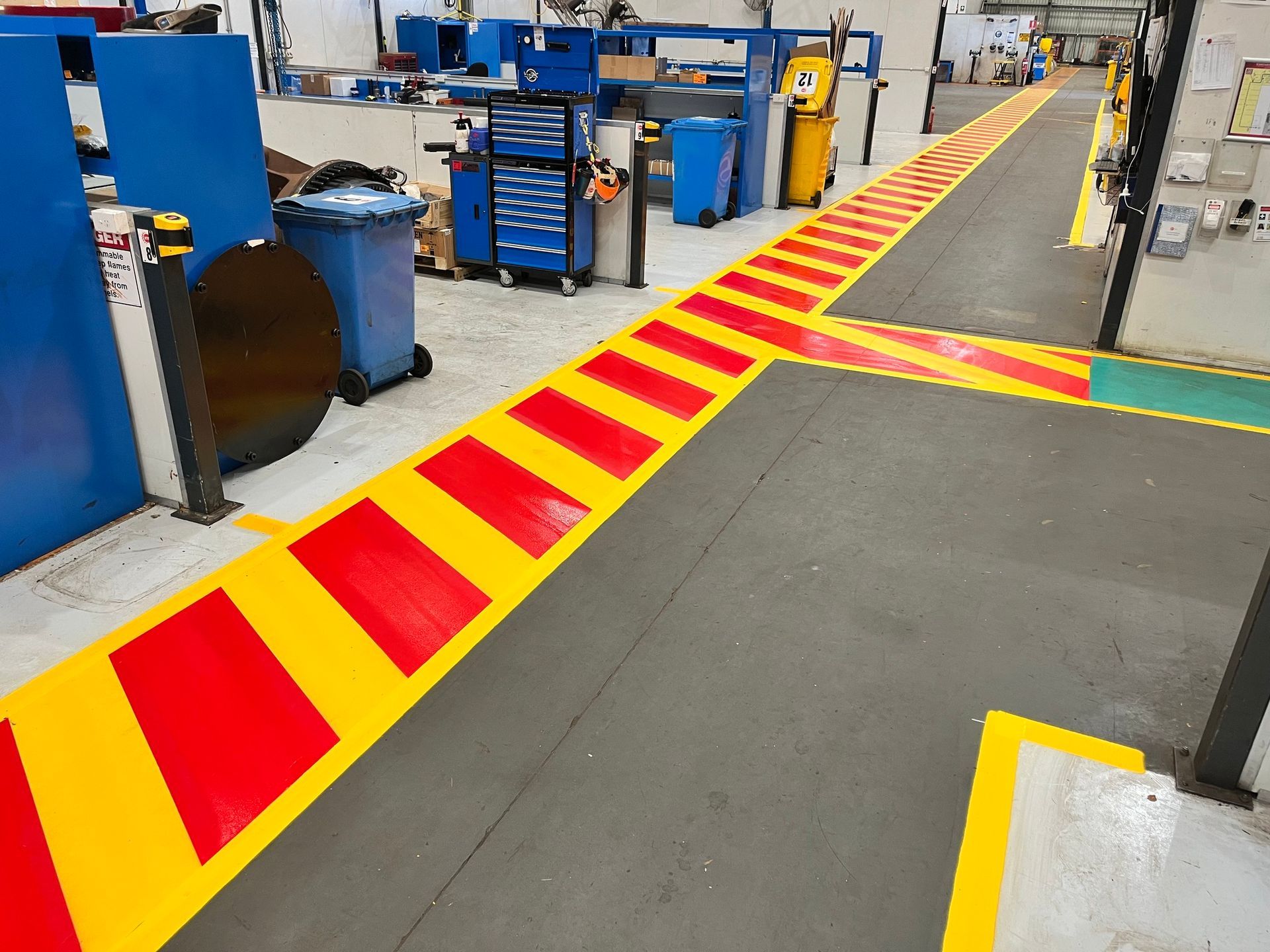 Factory floor with red and yellow painted hazard markings. Grey and blue machinery and equipment in the background.