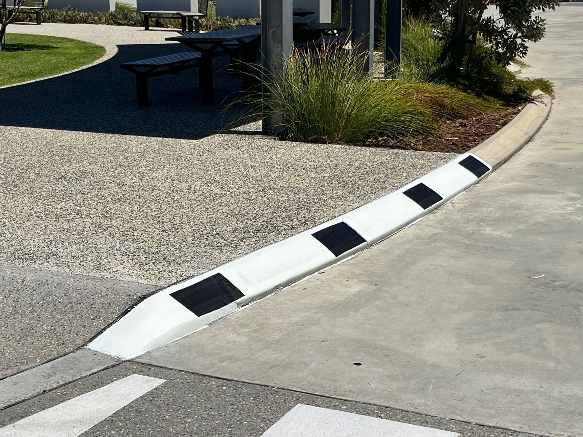 Concrete curb with alternating black and white squares, bordering a pebbled pathway and garden.
