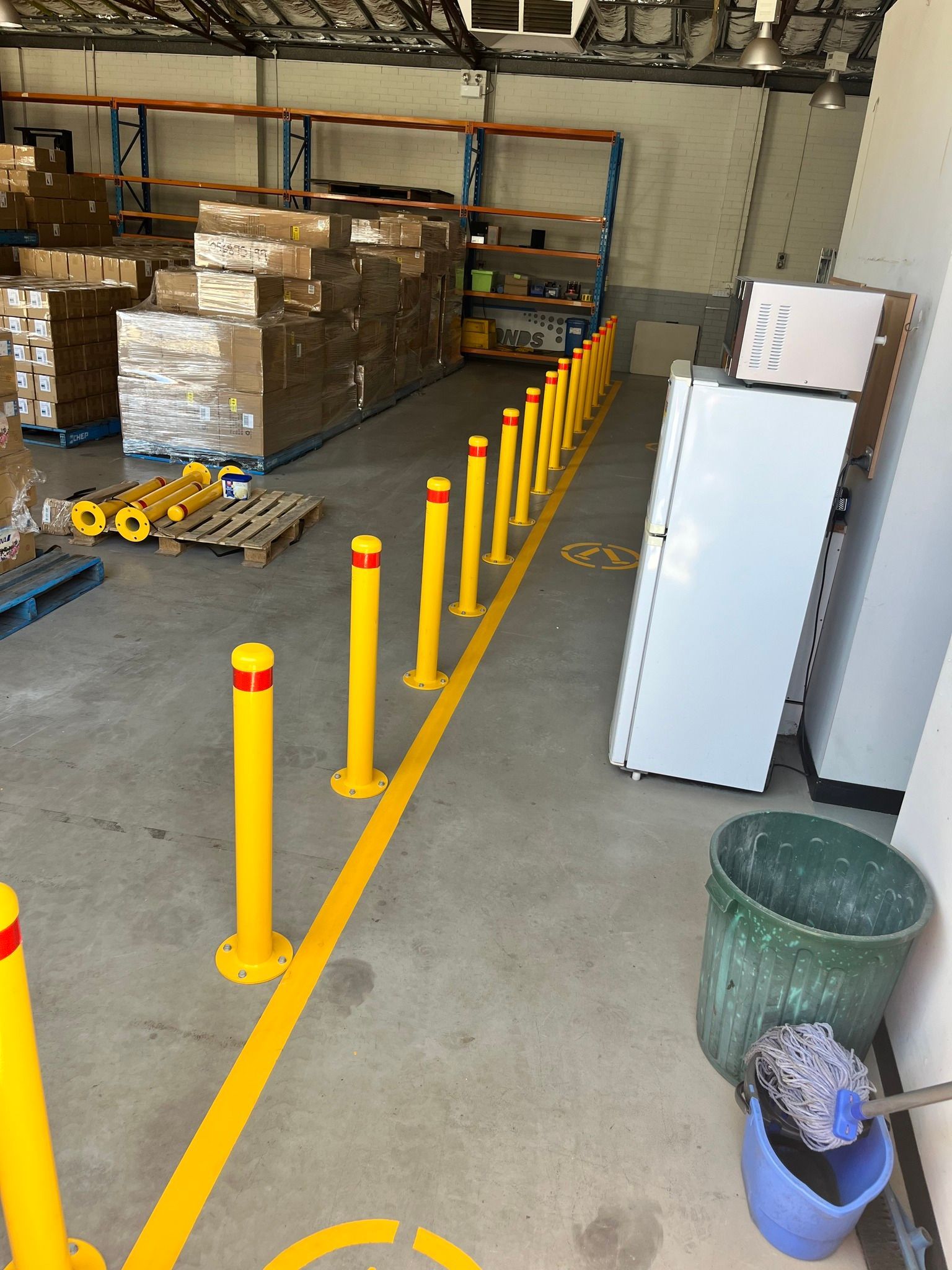 Yellow bollards line a warehouse walkway. Pallets, shelving, and boxes are visible.