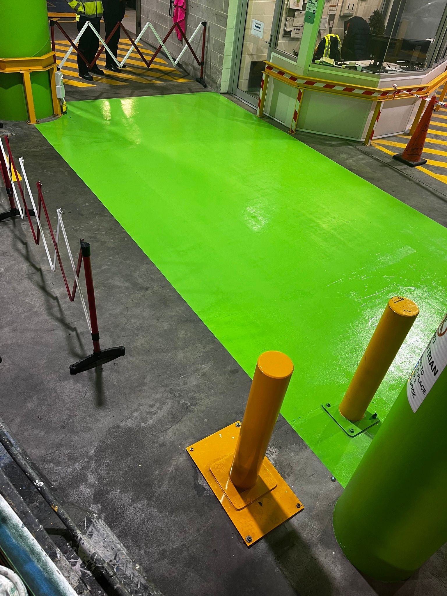 Bright green painted pathway in industrial setting, orange bollards, folding barrier, and workers in background.