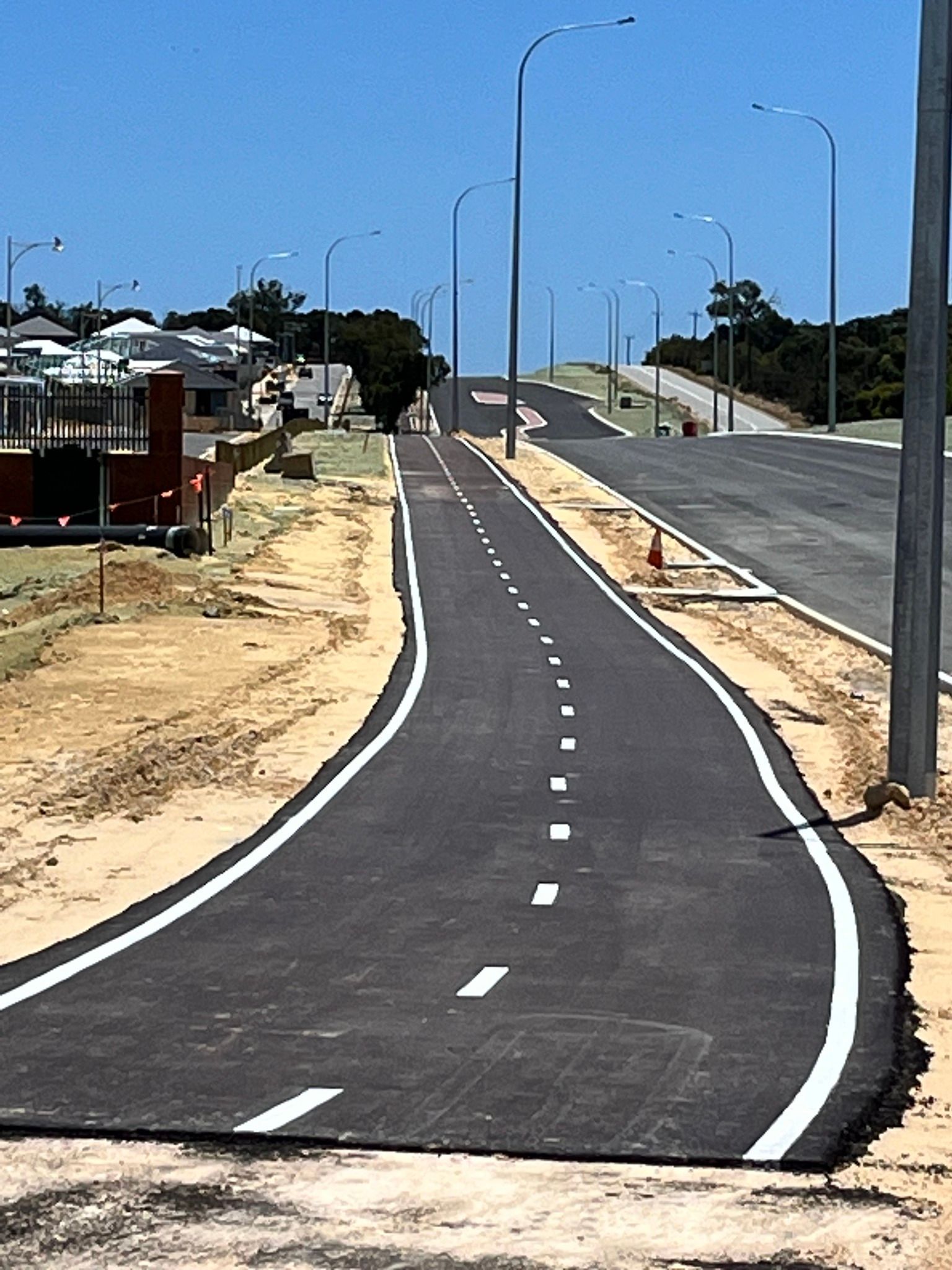 Paved road curving uphill with lane markings, flanked by dirt and tall streetlights under a blue sky.