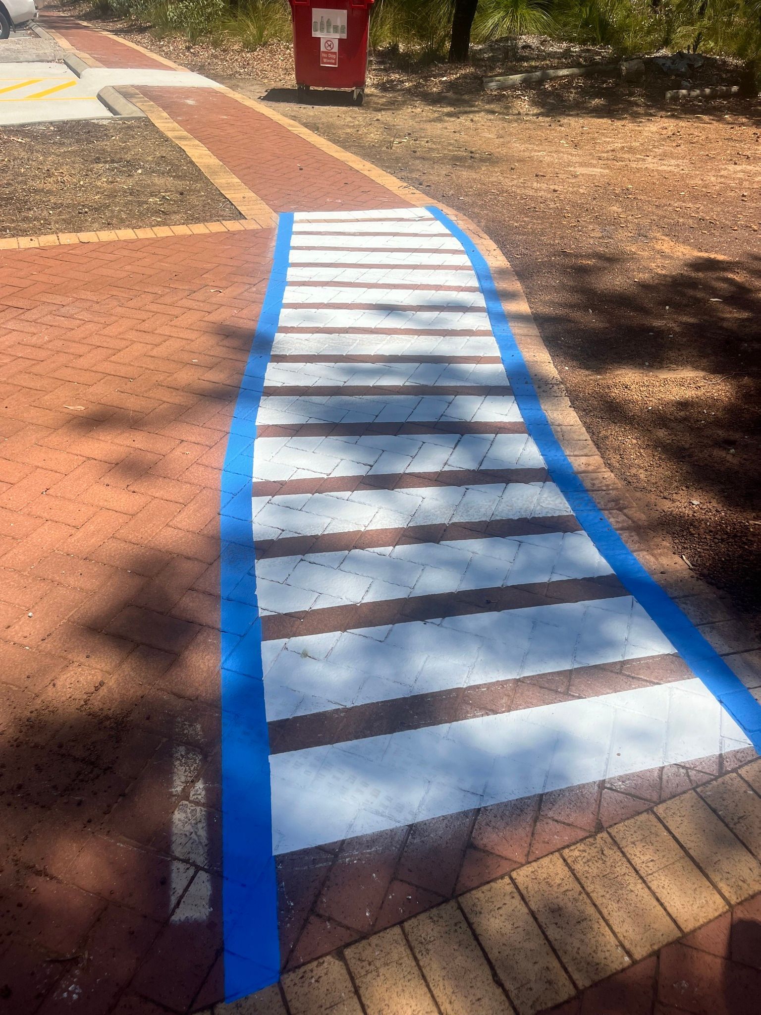 Crosswalk painted on brick path, outlined in blue, leading to a path with a red trash can in the background.
