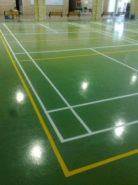 Green badminton court with white and yellow lines, inside a gymnasium.