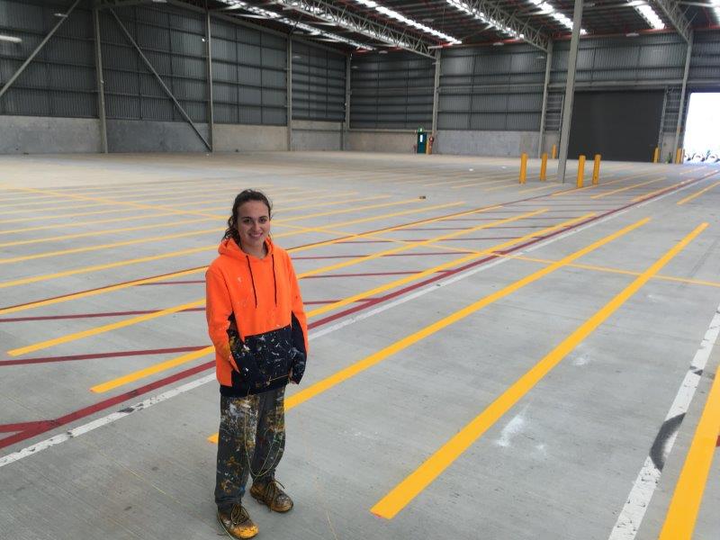 Woman in orange vest stands in a large warehouse with newly painted yellow floor markings.