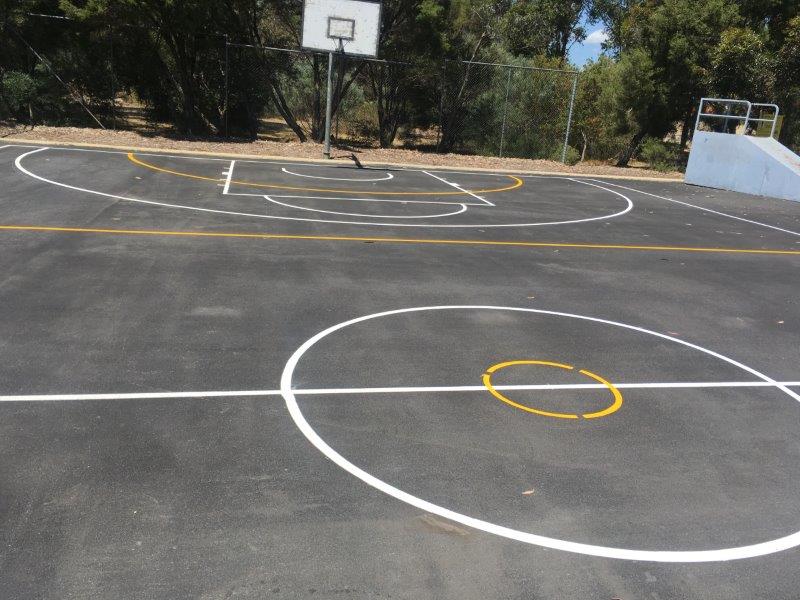 Basketball court with white and yellow lines, a hoop, and trees in the background.