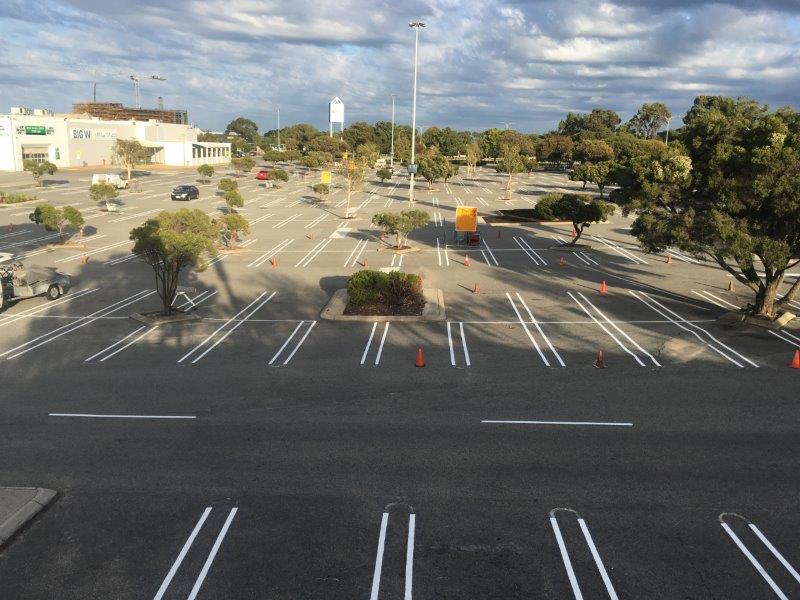 Empty parking lot with white-painted lines and small trees.  A few cars are parked. The sky is partly cloudy.