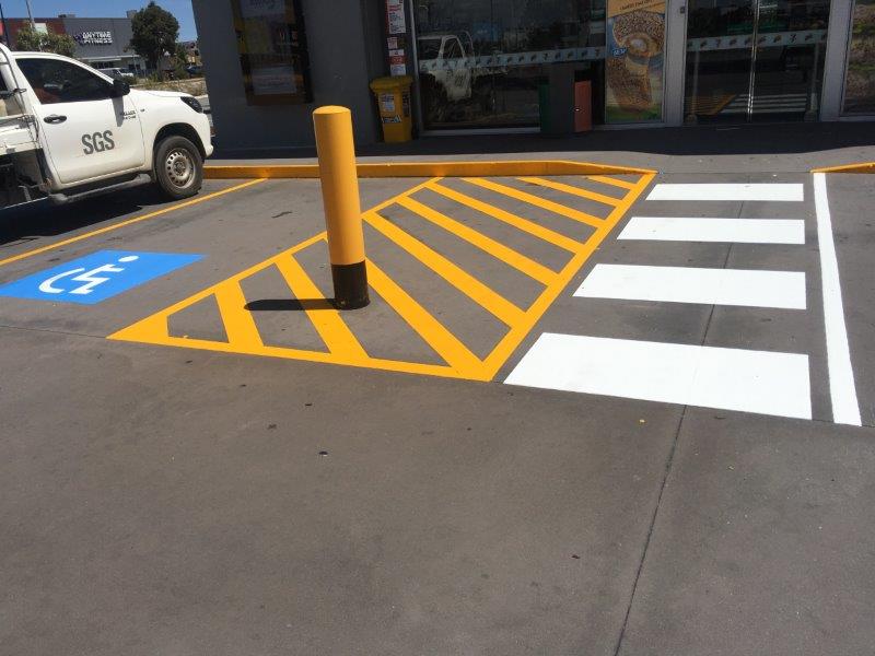 Crosswalk and disabled parking space painted on asphalt near a building entrance with a yellow bollard.
