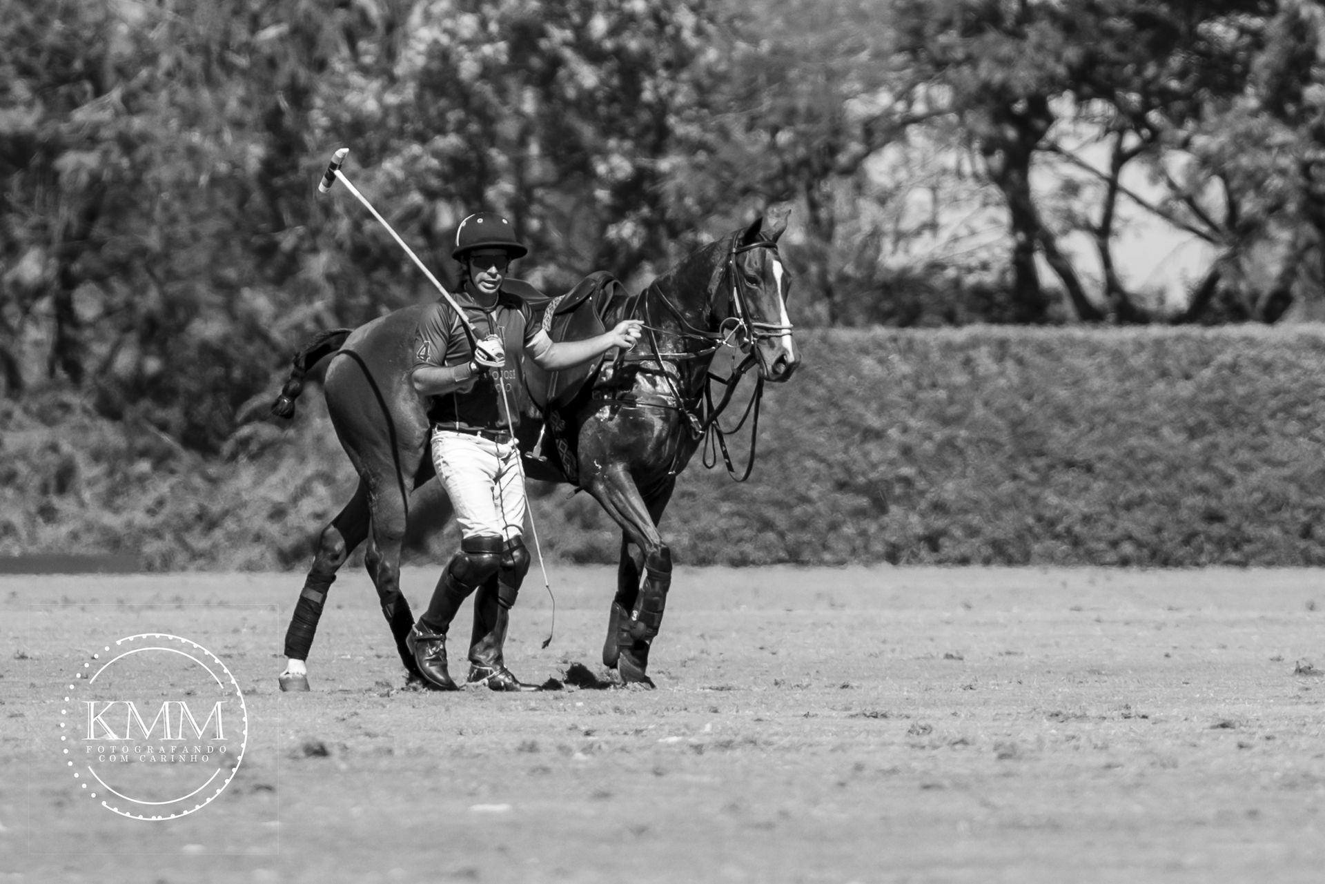 Polo player in helmet, black boots, and white pants leads horse on a grassy field.