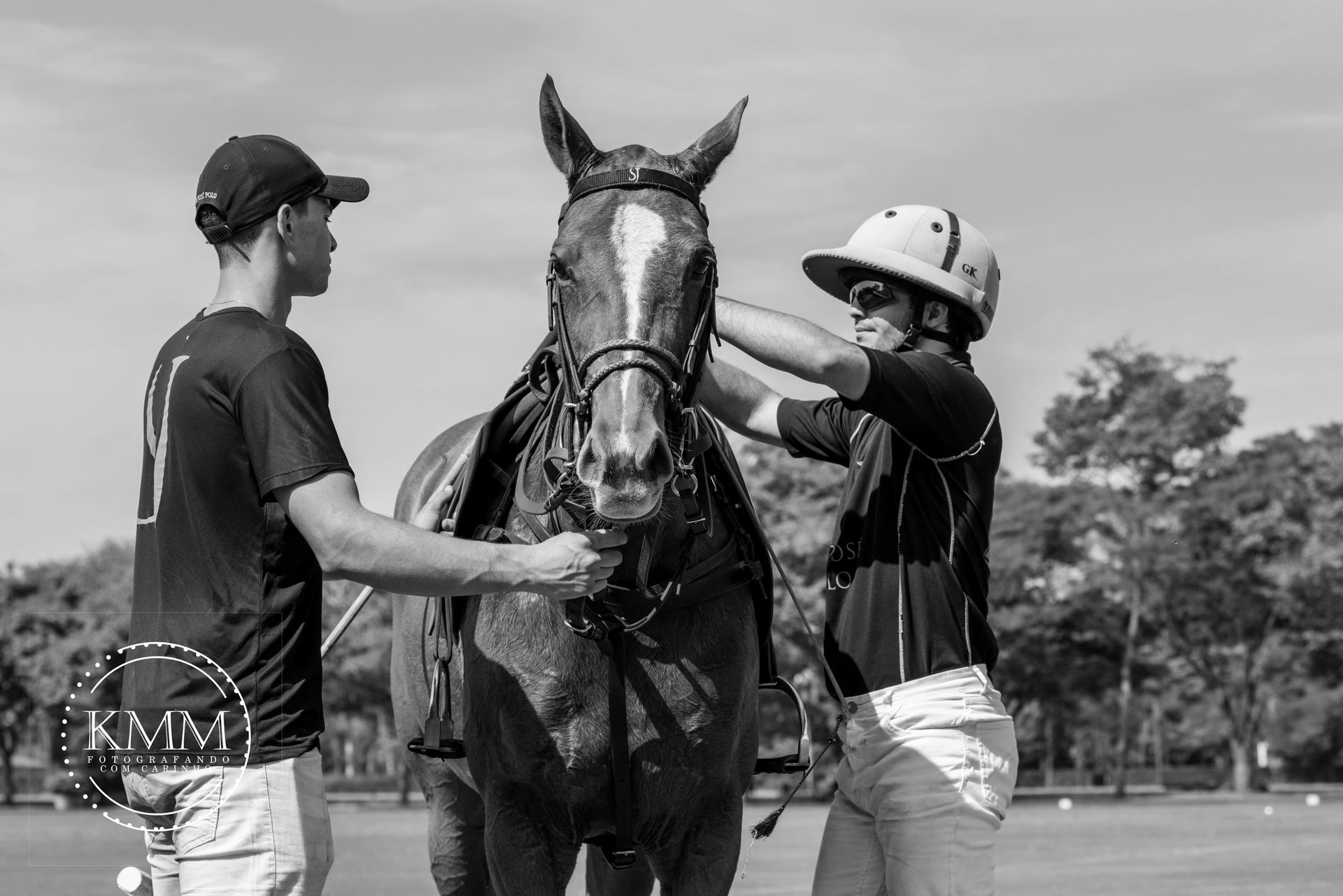 Two men preparing a horse for polo. One adjusts the bridle, the other holds the reins. Outdoors, sunny day.