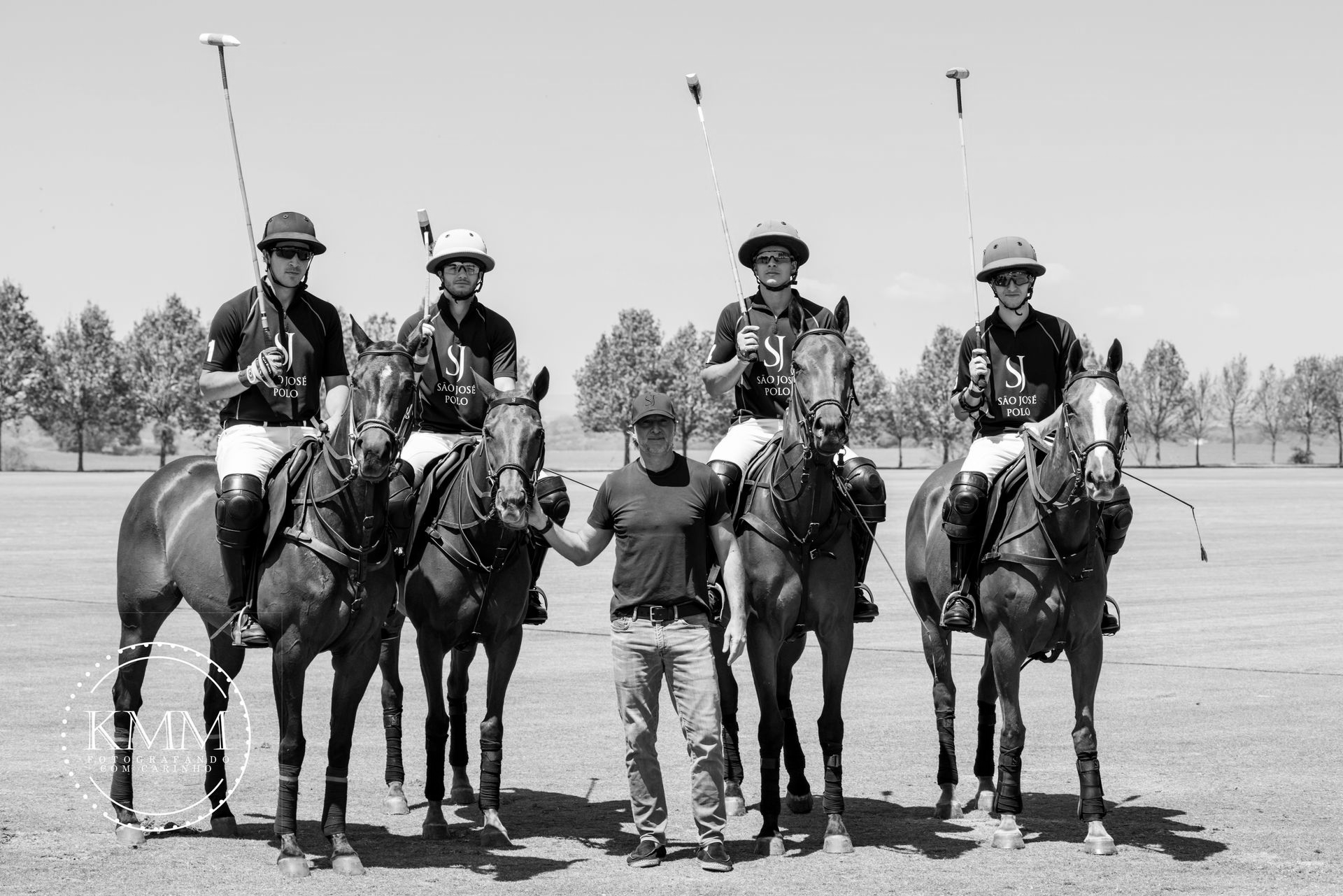 Polo team on horseback, posed on a field with a coach in the center. Sunlit day.