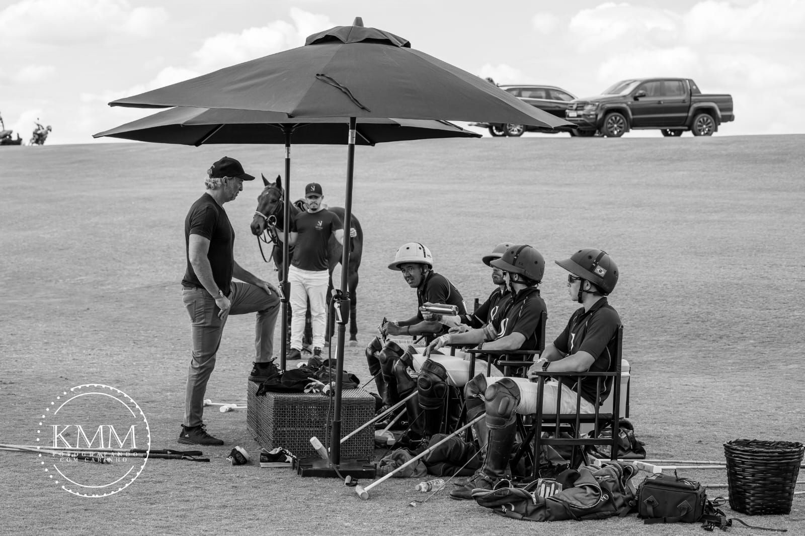 Polo players and staff under a large umbrella. Horses, equipment, and vehicles on a grassy field.