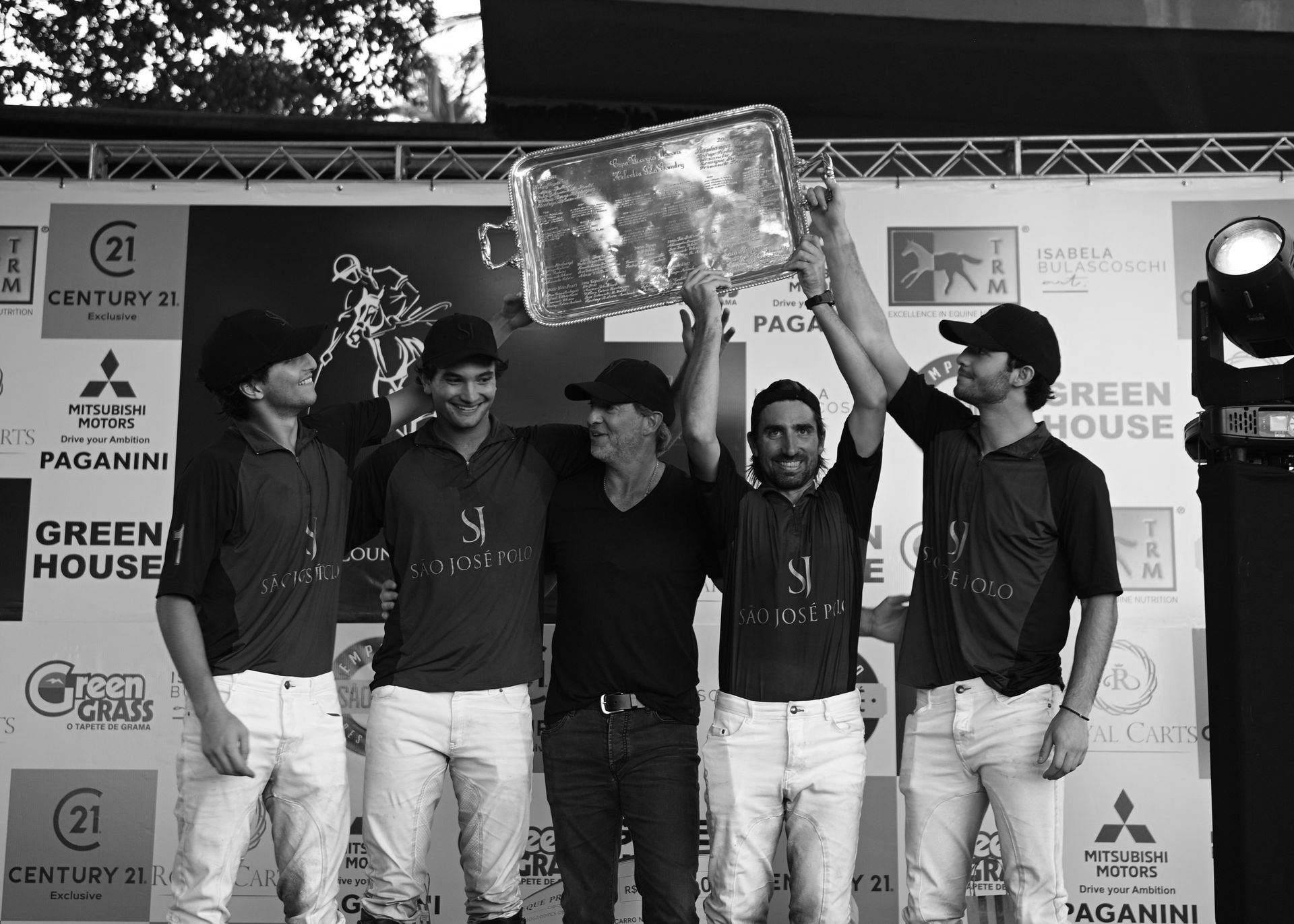 Five people on stage raising a trophy, wearing brown shirts and white pants, smiling at a competition event.