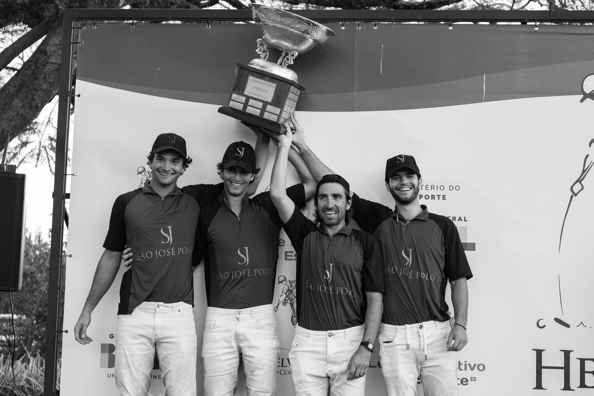 Four teammates in matching shirts and white pants hold up a trophy on a stage backdrop.