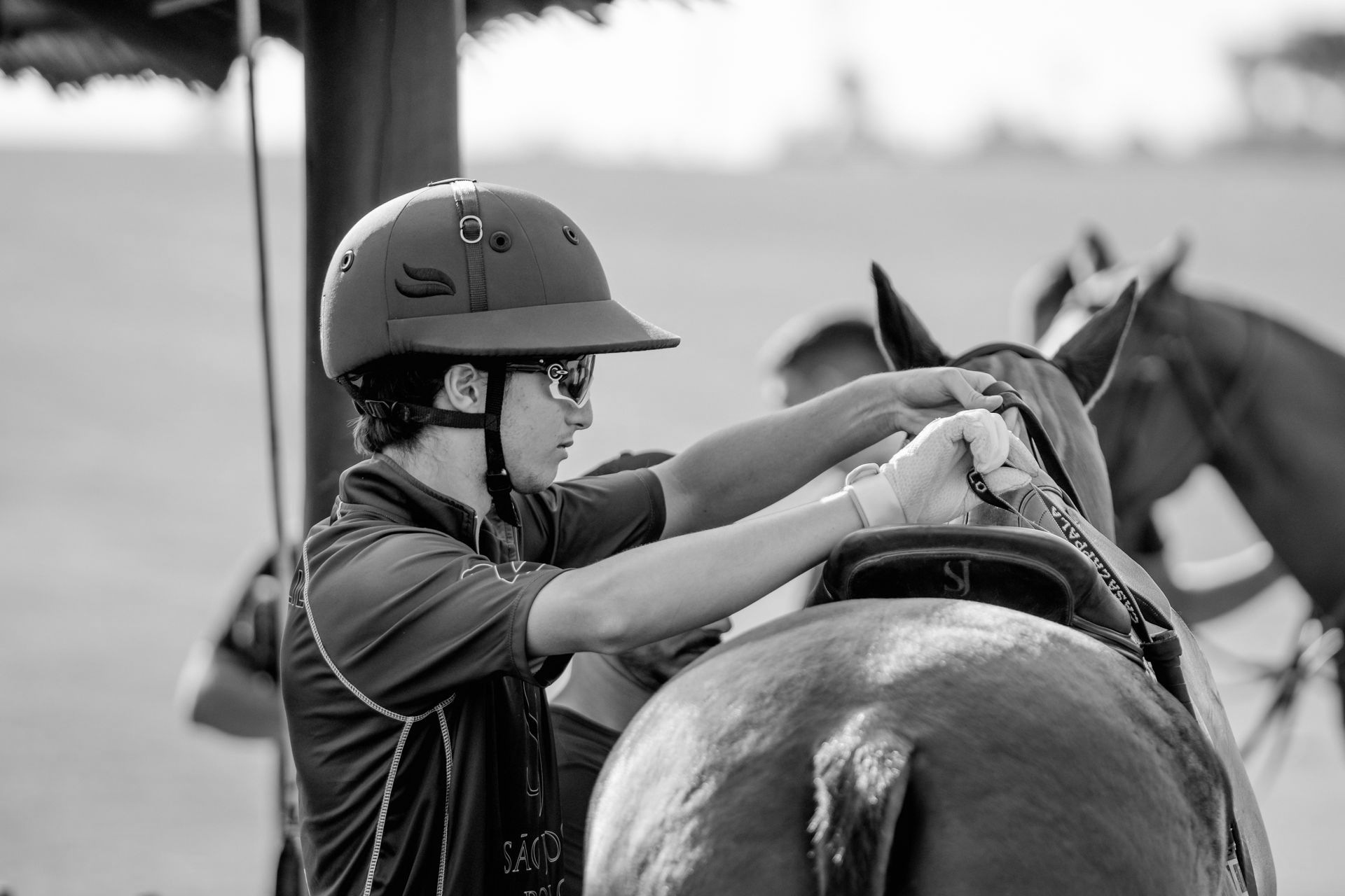 Person in helmet adjusts saddle on a horse in an outdoor setting.