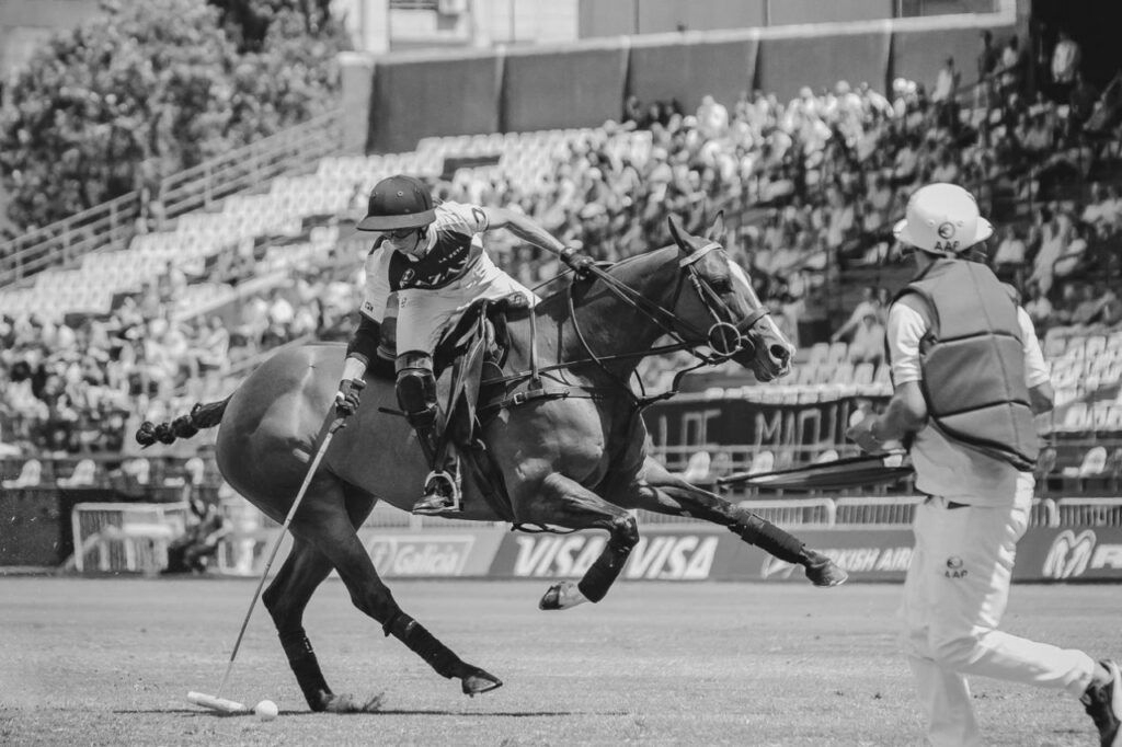 A polo player on horseback, losing balance. A referee runs nearby, stadium in background.