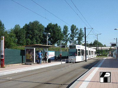 Eine weiße Straßenbahn an einer Haltestelle mit wartenden Menschen. Oberleitungen, Unterstand und Grünflächen sind sichtbar.