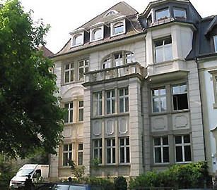 Verziertes Steingebäude mit mehreren Fenstern und einem Balkon; im Vordergrund sind ein Lieferwagen und Bäume zu sehen.
