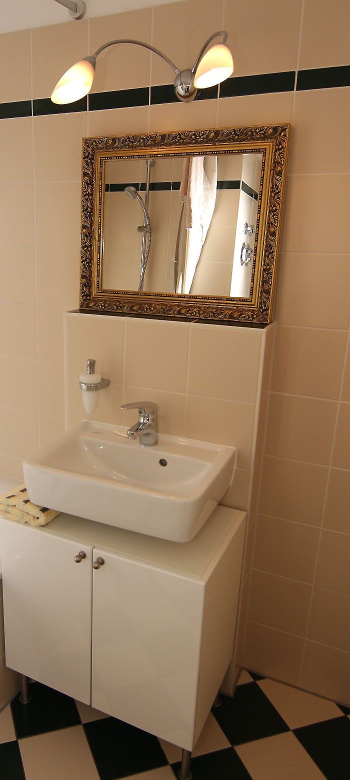 Waschbecken mit goldgerahmtem Spiegel, darüber weißer Schrank, schwarz-weiß karierter Boden.