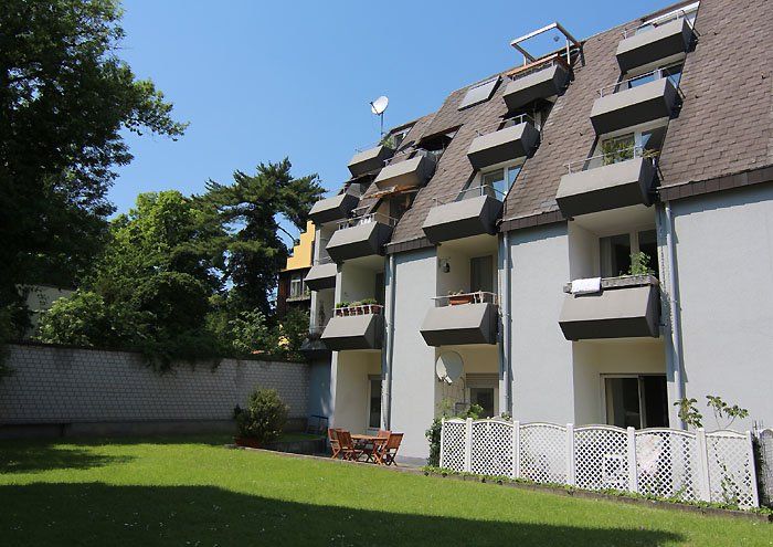 Mehrfamilienhaus mit vielen Balkonen, grünem Rasen und Bäumen an einem sonnigen Tag.