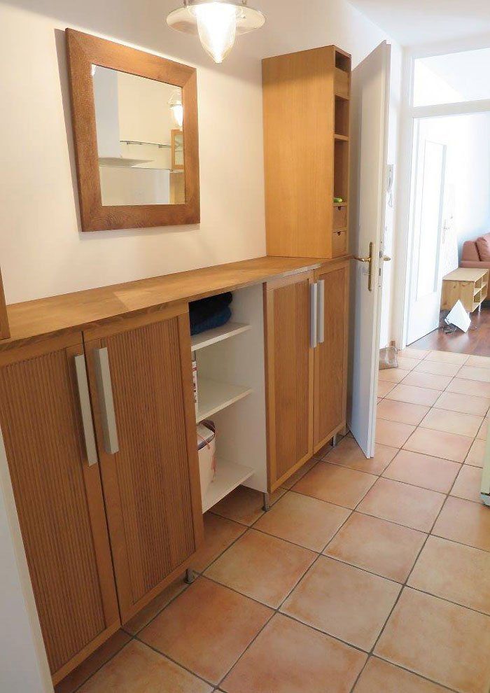 Flur mit hellen Holzschränken, Spiegel und offener Tür zu einem weiteren Raum. Terrakottafliesenboden.