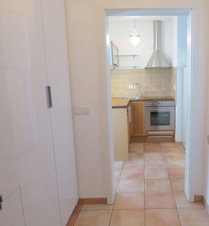 Schmaler Flur, der zu einer Küche mit hellen Holzschränken, Geräten aus Edelstahl und Fliesenboden führt.