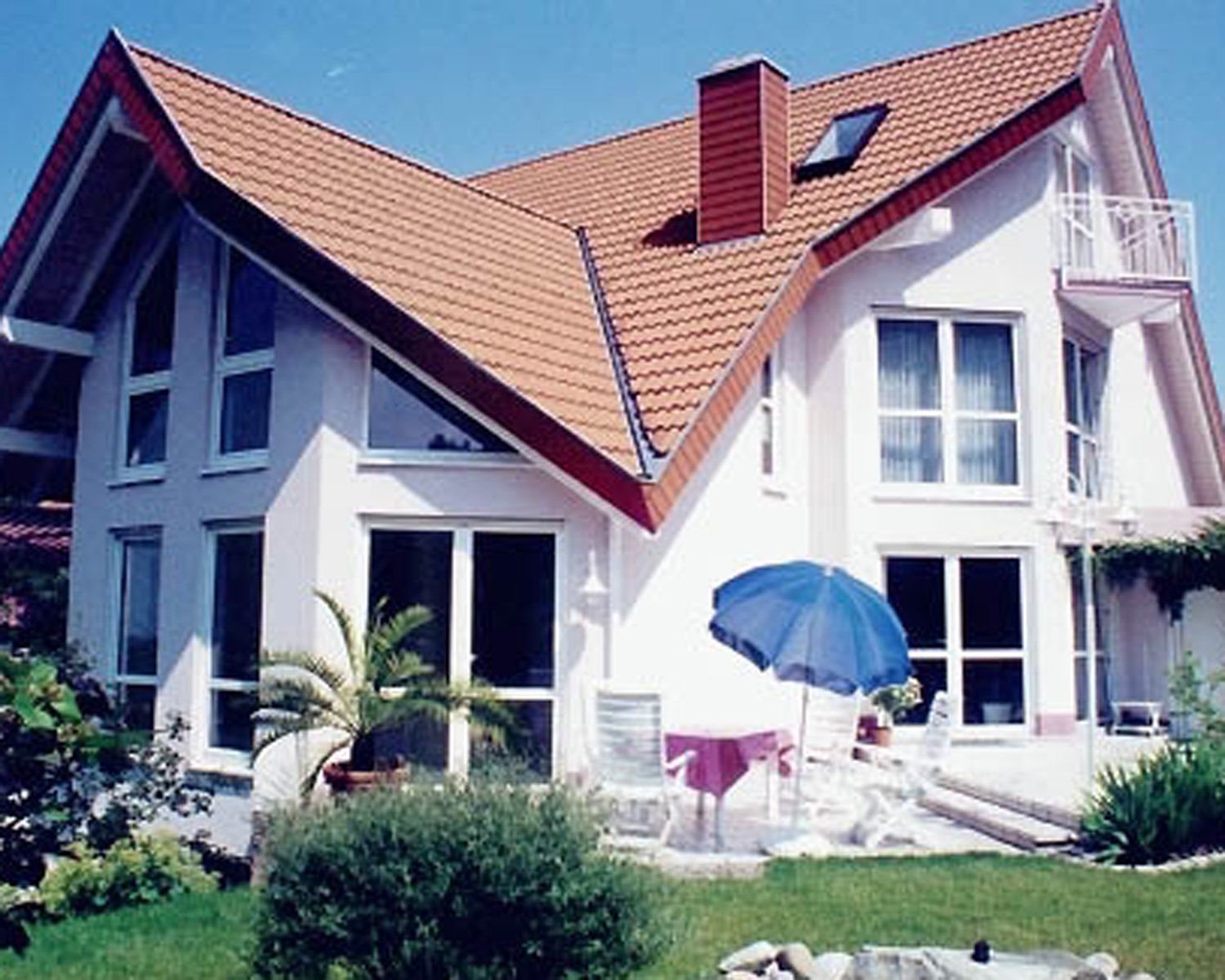 Zweistöckiges weißes Haus mit rotem Dach und Schornstein, blauer Sonnenschirm auf der Terrasse vor dem Haus.
