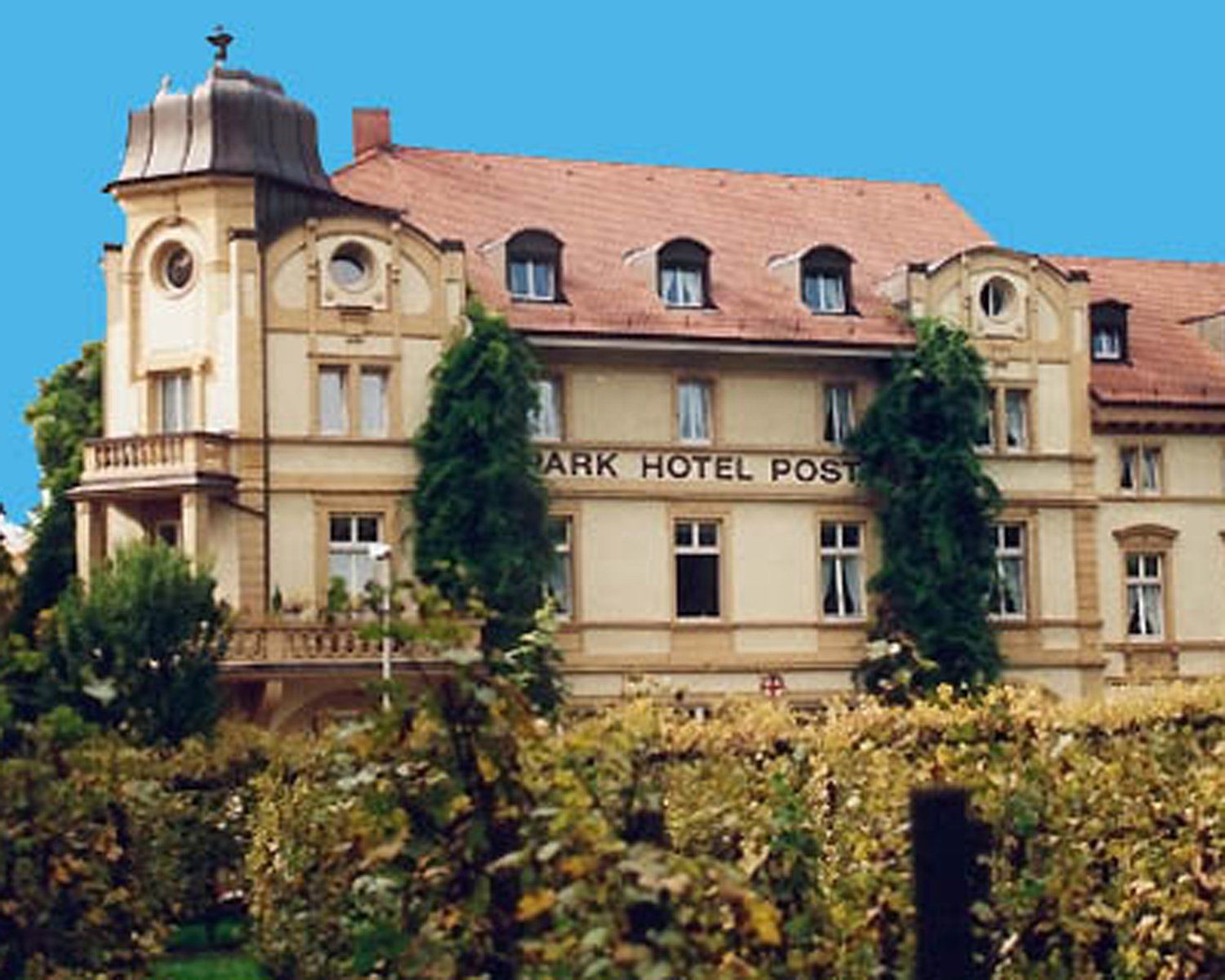 Park Hotel Post, ein mehrstöckiges hellbraunes Gebäude mit rotem Dach, in der Nähe von Weinreben.