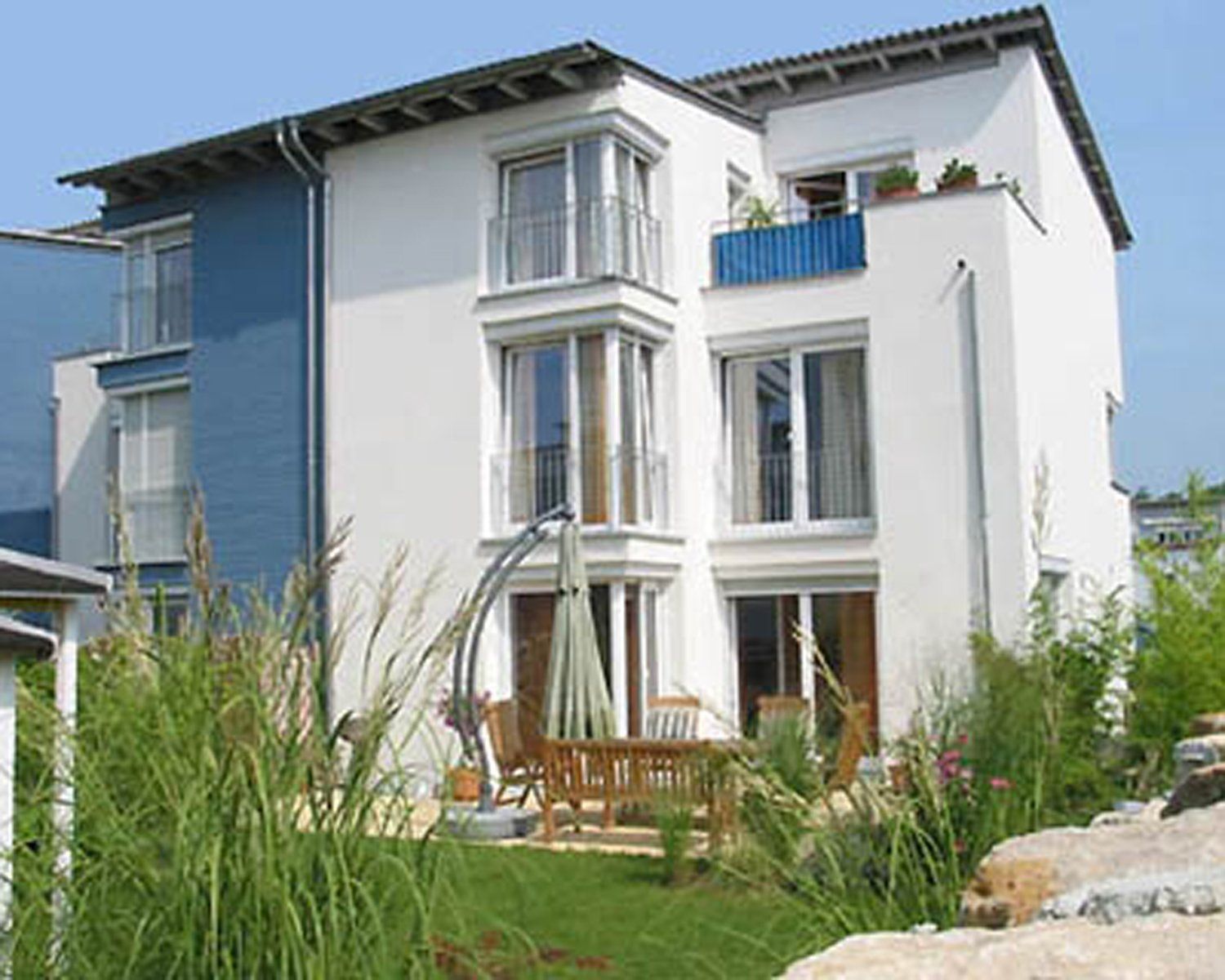 Modernes weiß-blaues Haus mit Balkonen und Sitzgelegenheiten im Freien. Grünes Gras und Ziergräser im Vordergrund.