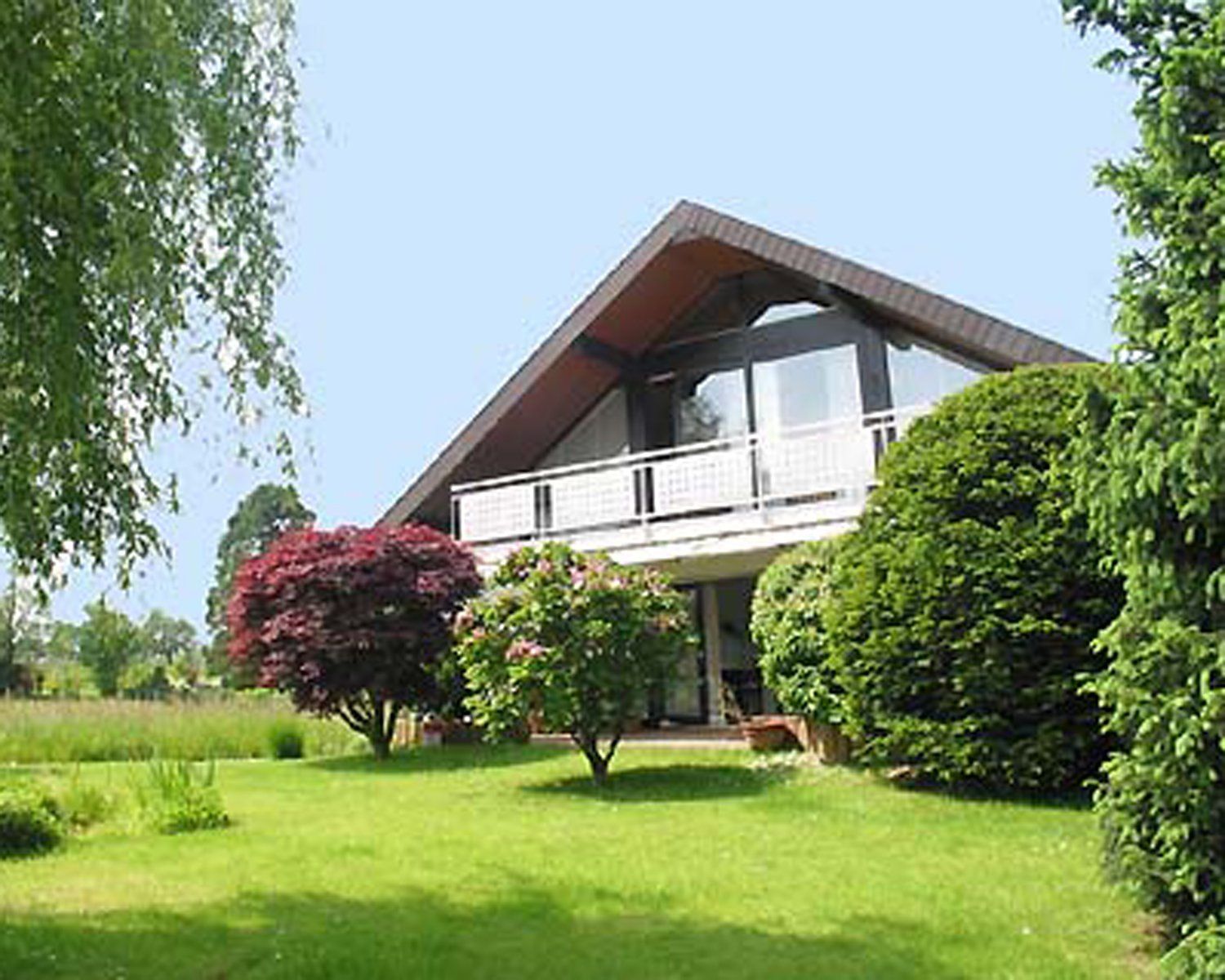Ein modernes Haus mit Schrägdach und Balkon, umgeben von grünem Rasen und Bäumen.