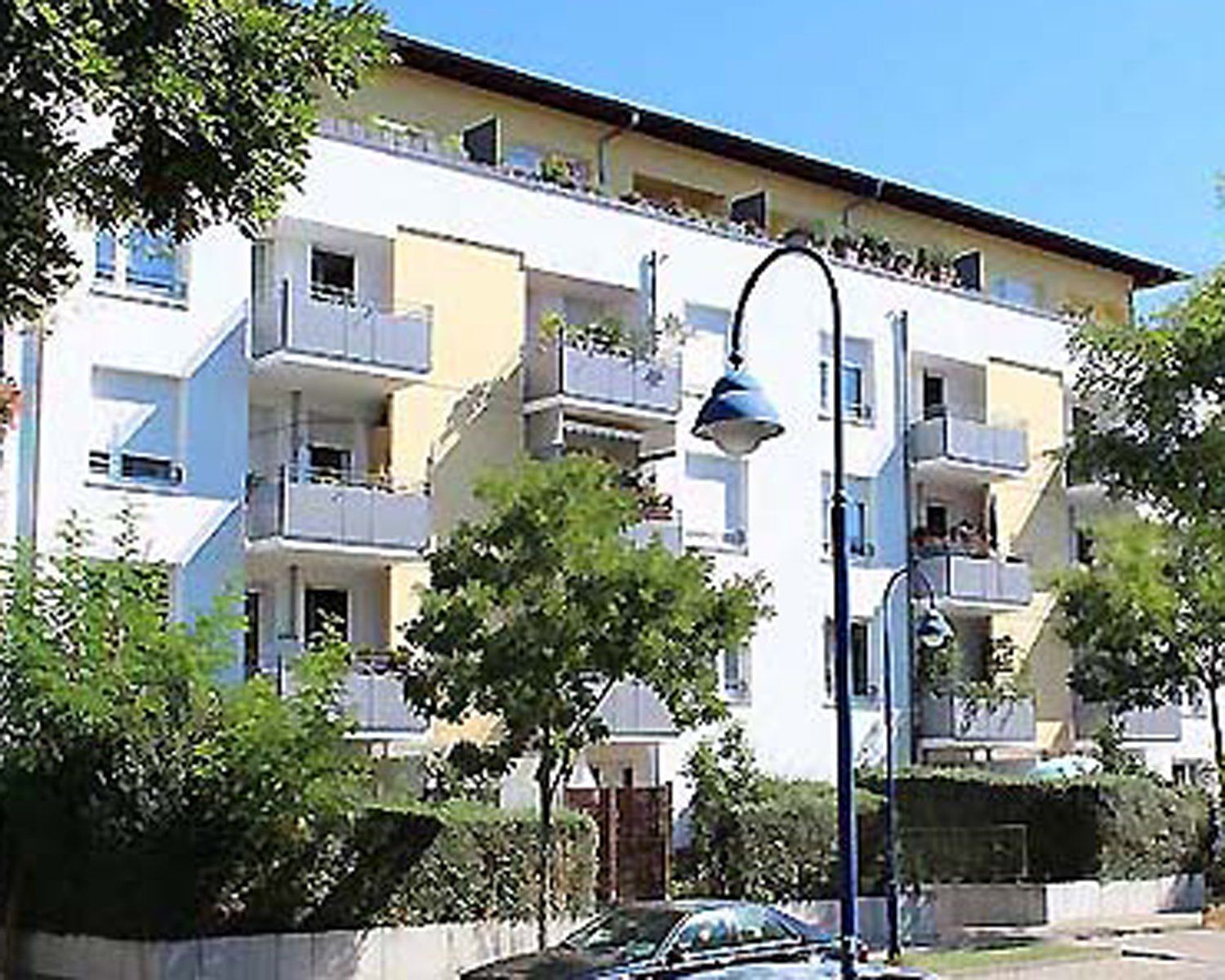 Mehrfamilienhaus mit Balkonen, weiß-gelber Fassade, Bäumen und einer Straßenlaterne im Vordergrund.