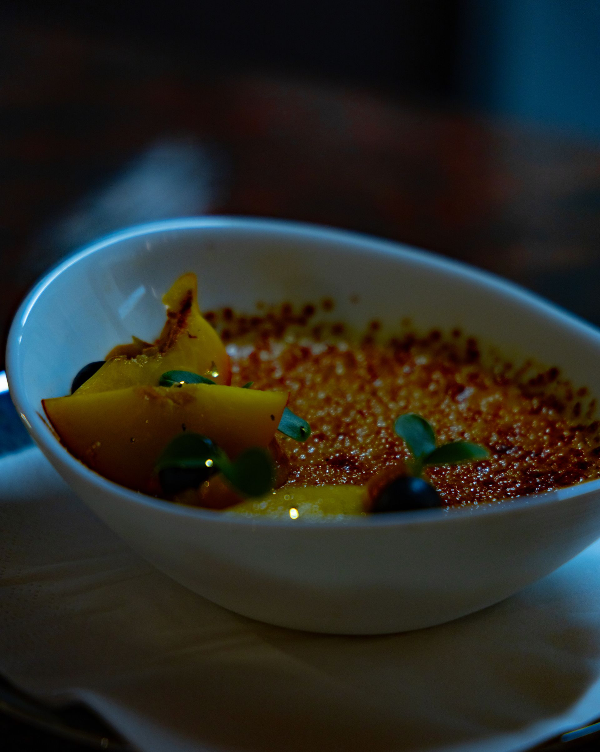 Crème brûlée-Dessert in einer weißen Schüssel, garniert mit gelben Früchten und grünen Kräuterzweigen.