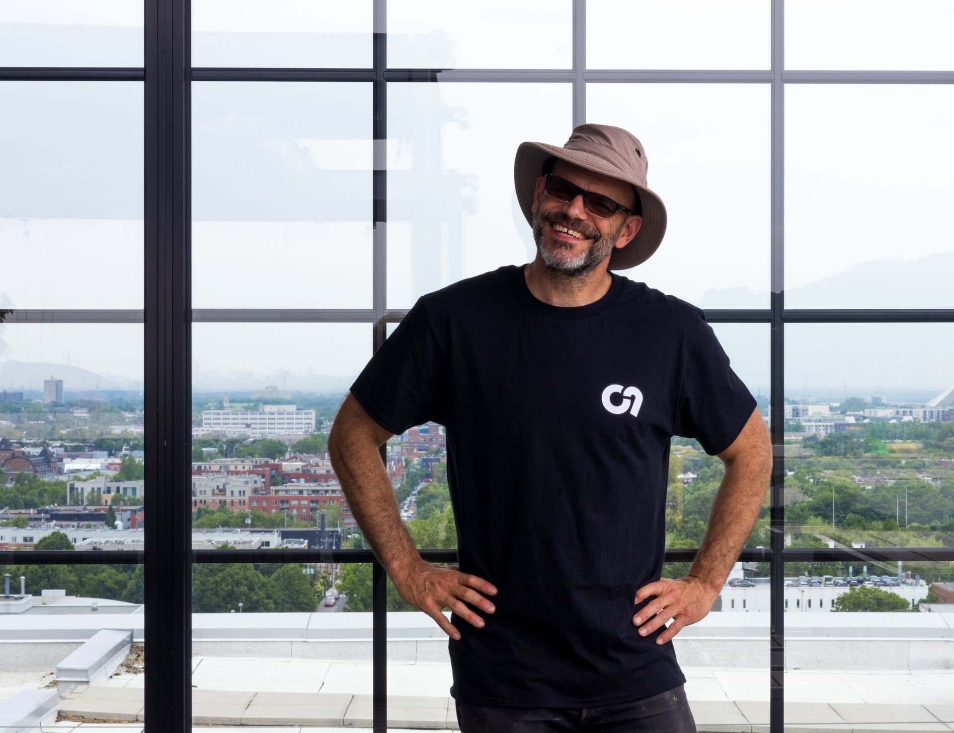 Un homme avec un chapeau et des lunettes de soleil sourit, les mains sur les hanches, devant une fenêtre avec vue sur la ville.