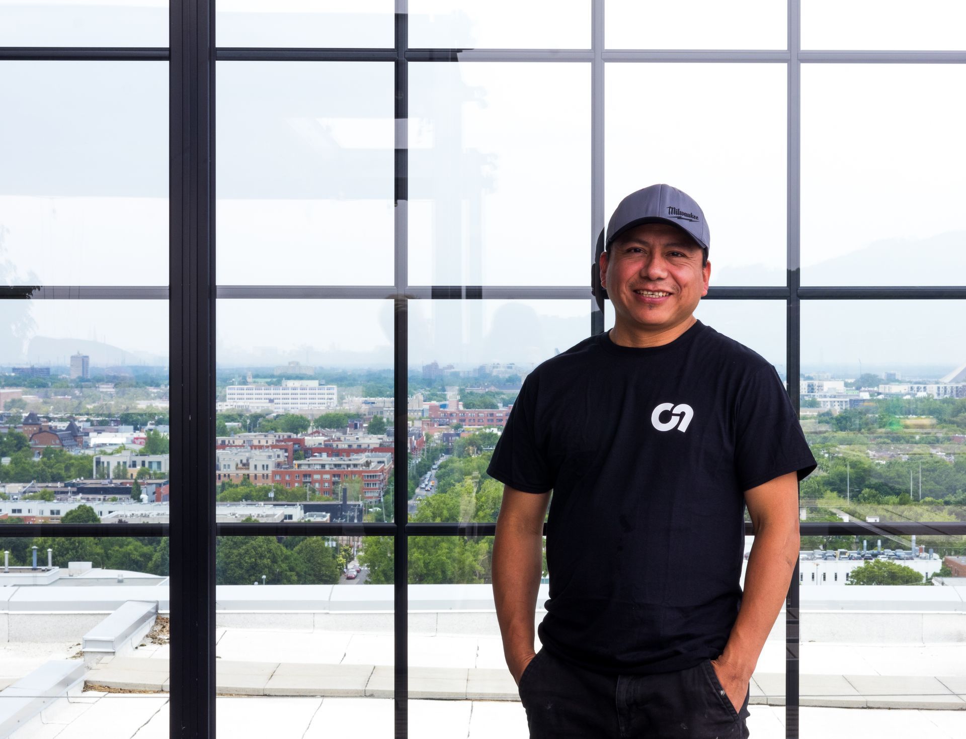 Homme avec casquette et chemise noire, debout devant une fenêtre avec vue sur la ville, les mains dans les poches, souriant.
