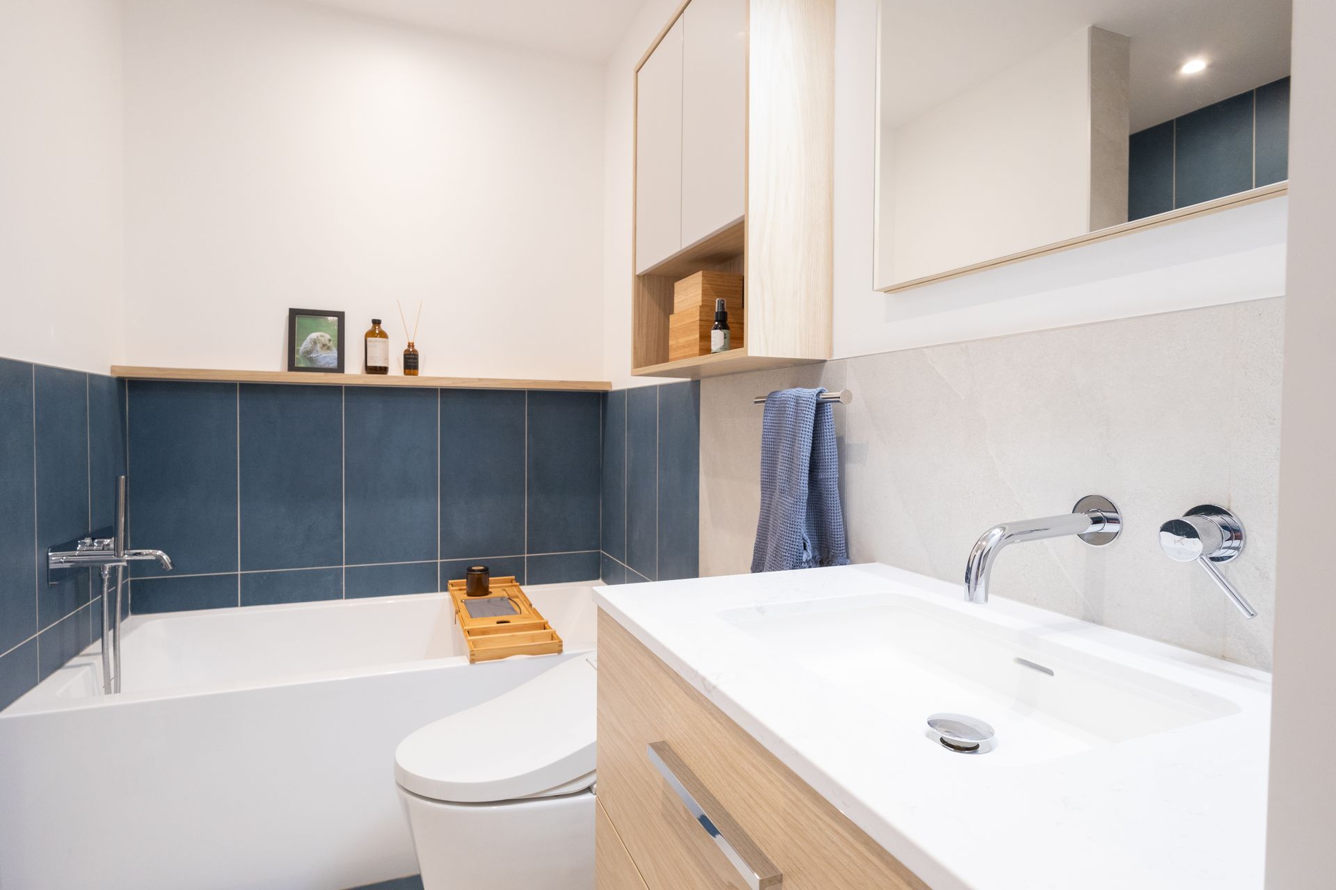 Salle de bain moderne avec murs blancs, carrelage bleu et meubles en bois clair. Elle comprend un lavabo, une baignoire et des toilettes.