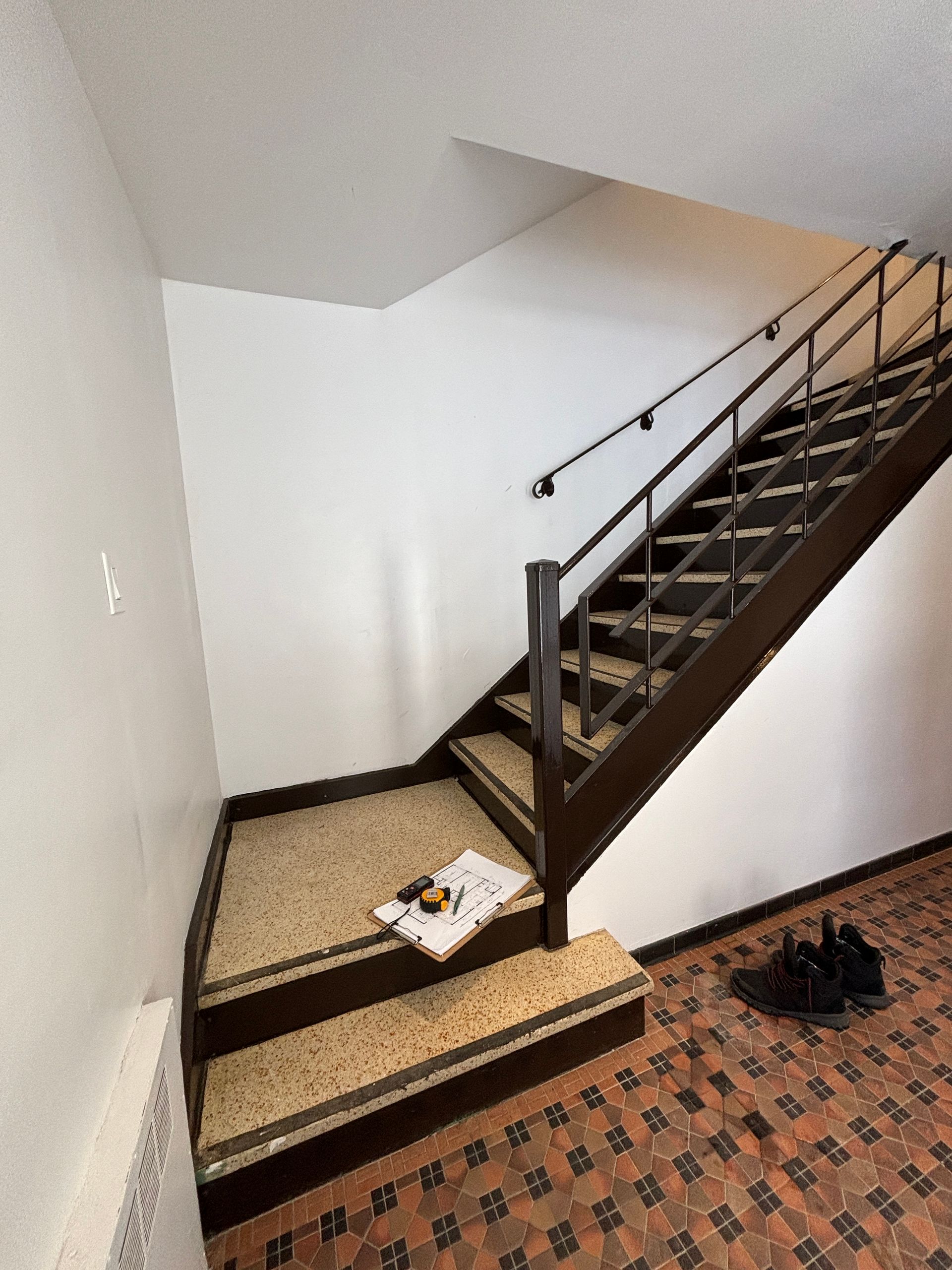 Escalier avec rampe et marches marron, sol à motifs et murs blancs. Papier et chaussures en bas.
